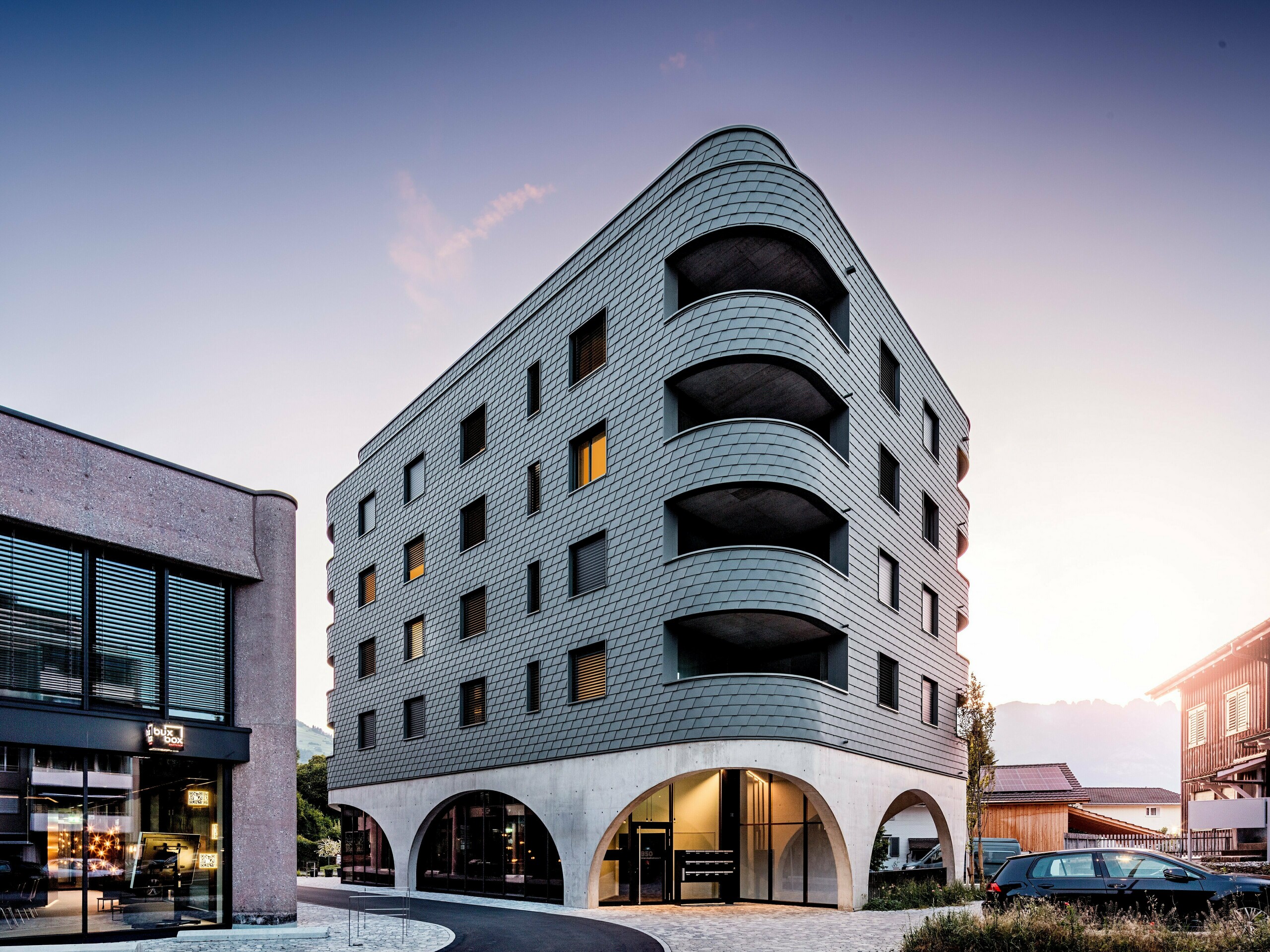 Moderná polyfunkčná budova v švajčiarskom meste Buchs s výrazným zaobleným trojuholníkovým tvarom a súvislou fasádou z PREFA šindľov v odtieni P.10 svetlošedá. Šupinovitý hliníkový plášť obklopuje budovu ako plazová štruktúra a zdôrazňuje plynulé zaoblenia a hlboko zasadené balkóny. Nad masívnym podstavcom s elegantnými betónovými arkádami pôsobí ľahká, odolná proti poveternostným vplyvom fasáda PREFA obzvlášť výrazne. Presne položené hliníkové šindle zaisťujú homogénny, trvanlivý a nenáročný na údržbu plášť budovy s vysokou rozpoznateľnosťou.