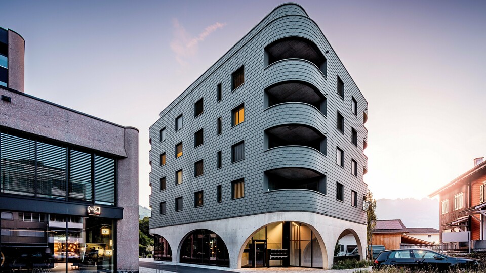 Moderná polyfunkčná budova v švajčiarskom meste Buchs s výrazným zaobleným trojuholníkovým tvarom a súvislou fasádou z PREFA šindľov v odtieni P.10 svetlošedá. Šupinovitý hliníkový plášť obklopuje budovu ako plazová štruktúra a zdôrazňuje plynulé zaoblenia a hlboko zasadené balkóny. Nad masívnym podstavcom s elegantnými betónovými arkádami pôsobí ľahká, odolná proti poveternostným vplyvom fasáda PREFA obzvlášť výrazne. Presne položené hliníkové šindle zaisťujú homogénny, trvanlivý a nenáročný na údržbu plášť budovy s vysokou rozpoznateľnosťou.