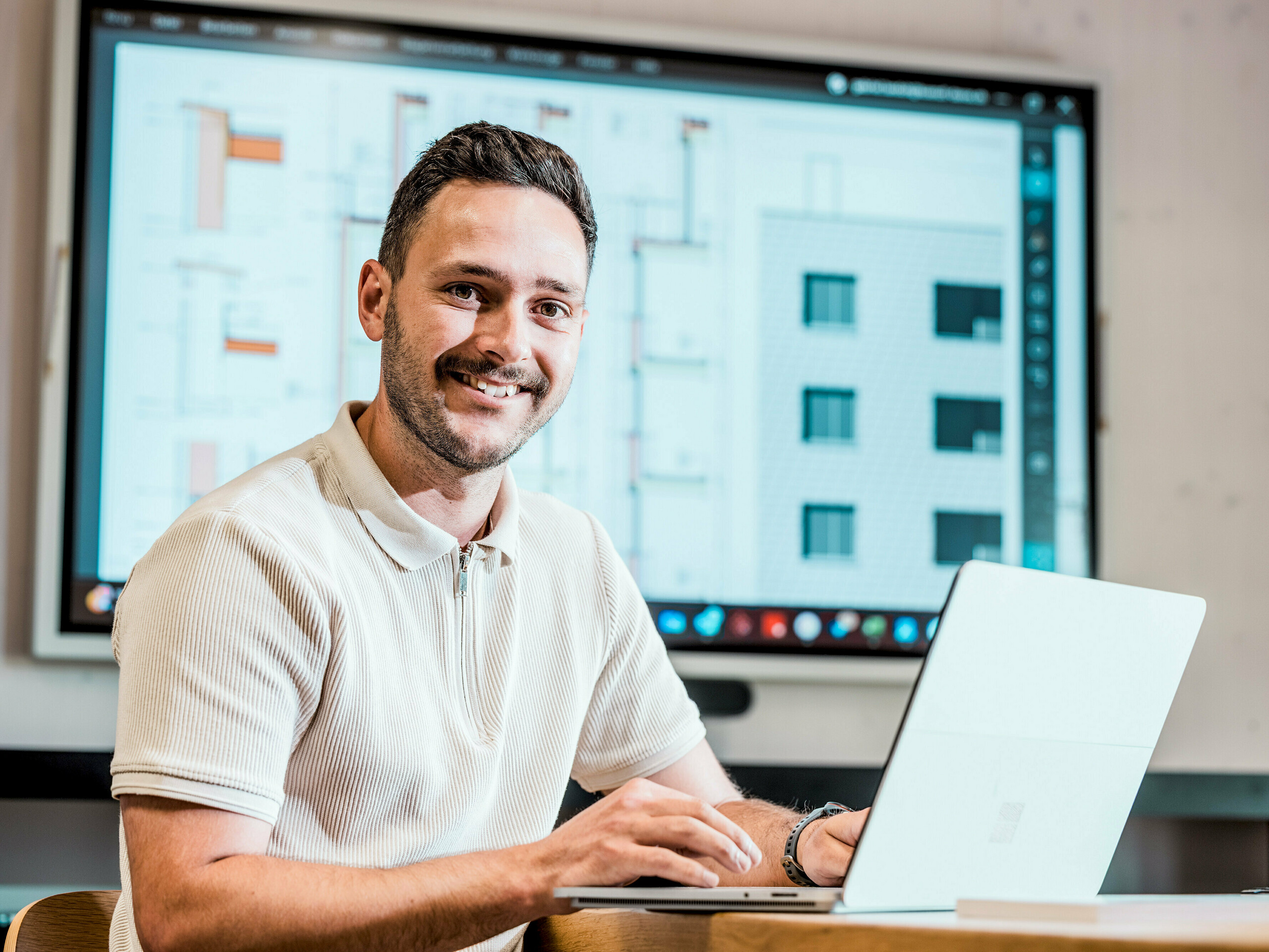 Portrét Gabriela Landolta, zamestnanca spoločnosti Künzli Davos AG, pri notebooku pred veľkou obrazovkou, na ktorej sú zobrazené digitálne stavebné plány a detaily fasády. Na monitore sú zobrazené technické výkresy a pohľady na modernú budovu so šupinovou fasádou, ktorá bola realizovaná s použitím PREFA fasádnych šindľov v farbe P.10 svetlošedá. Inovatívna hliníková fasáda presviedča svojou nízkou hmotnosťou, odolnosťou voči poveternostným vplyvom a dlhou životnosťou. Presné plánovanie a použitie vysoko kvalitných produktov PREFA sú základom pre tvarovo stabilný, nenáročný na údržbu plášť budovy s architektonickými nárokmi.