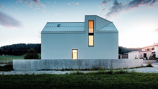 Obrázok ukazuje nápadný rodinný dom vo Freistadte, ktorý zaujme svojimi čistými líniami a minimalistickou architektúrou. Čisto biela fasáda PREFALZ z ohýbaného hliníka vytvára výraznú geometrickú štruktúru, ktorá dodáva budove moderný a nadčasový vzhľad. Strmá sedlová strecha s integrovaným vikierovým oknom a vysoké okná zaisťujú dostatok denného svetla a dodávajú interiéru otvorený a príjemný vzhľad. Použitie hliníka PREFALZ zaručuje nielen estetickú stálosť, ale aj vynikajúcu odolnosť voči poveternostným vplyvom a dlhú životnosť, čo robí dom funkčným a zároveň udržateľným. Čisté tvary a štýlové detaily prispievajú k elegantnému a zároveň praktickému riešeniu modernej obytnej architektúry.