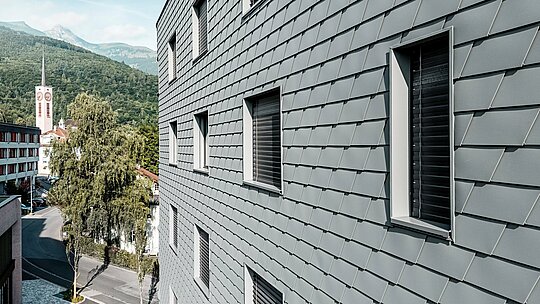 Detailný pohľad na fasádu domu Bernerhaus v Buchsi s presne položenými PREFA šindľami v odtieni P.10 svetlošedá. Šupinatý povrch hliníka vytvára jemný, živý vzor a zdôrazňuje jasne vyrezané okenné otvory s hlbokými osteniami. Ľahký, odolný voči poveternostným vplyvom hliník PREFA zaisťuje homogénny, tvarovo stabilný a nenáročný na údržbu plášť budovy. Pred alpskou kulisou sa vysokokvalitná kovová fasáda stáva moderným pútačom s nadčasovým vzhľadom.