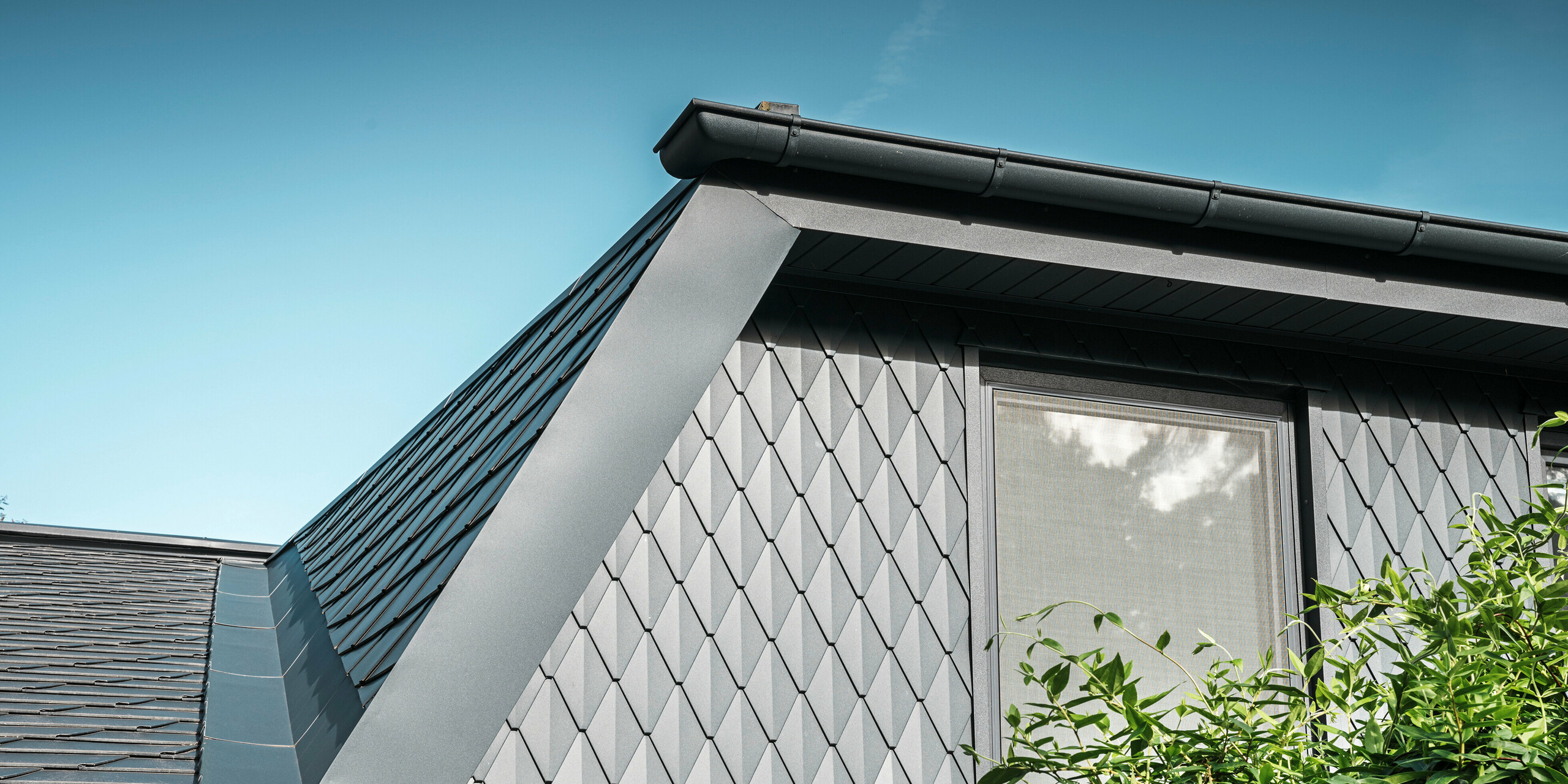 Trapézová vikierová strecha rodinného domu v Mechernichu, obložená malými strešnými šablónami PREFA v odtieni P.10 antracitová. Strecha je pokrytá strešnými šindľami PREFA DS.19 v rovnakom odtieni. Vysoko kvalitný hliníkový obklad zaisťuje odolnosť voči poveternostným vplyvom a moderný, estetický vzhľad.