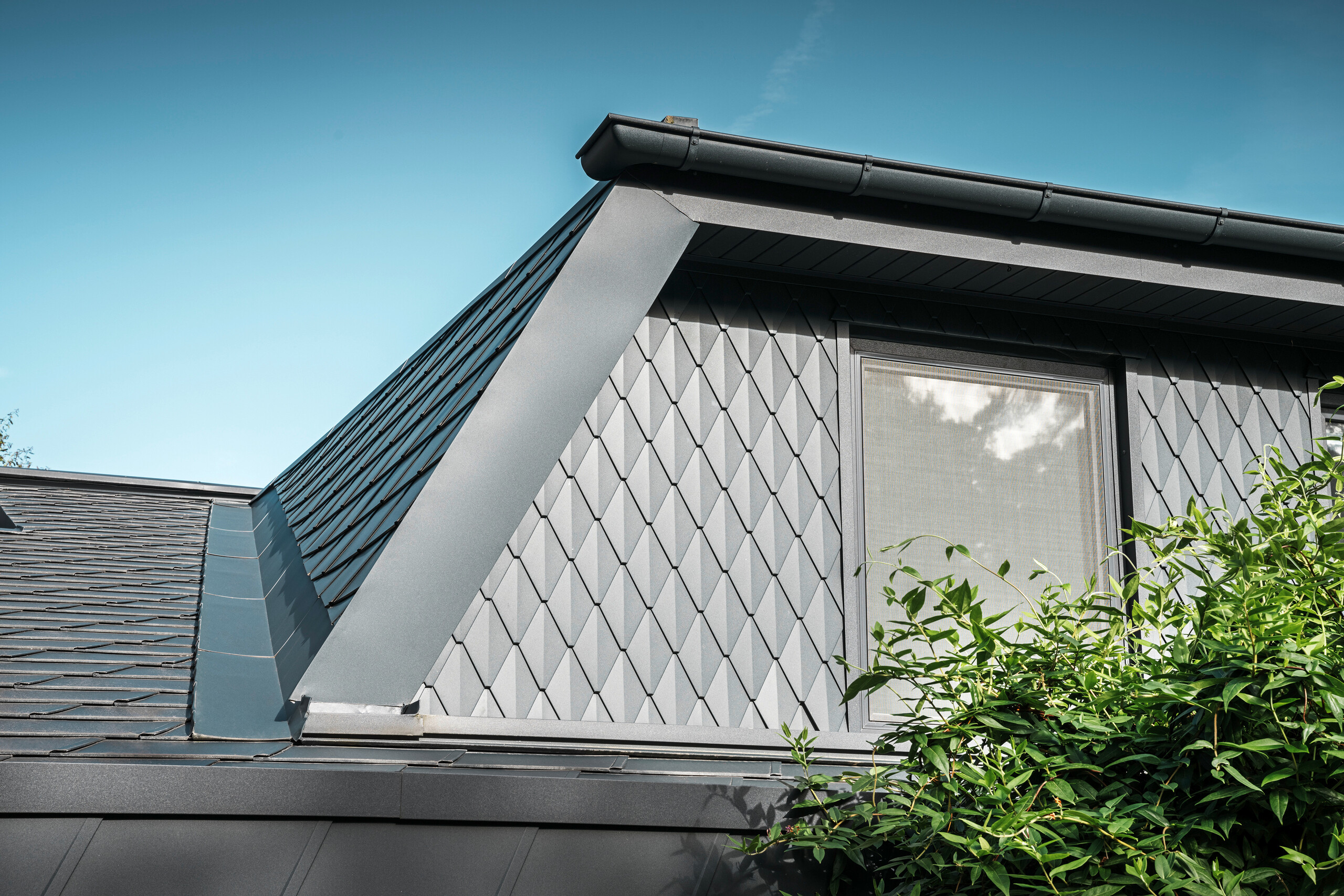 Trapézová vikierová strecha rodinného domu v Mechernichu, obložená malými strešnými šablónami PREFA v odtieni P.10 antracitová. Strecha je pokrytá strešnými šindľami PREFA DS.19 v rovnakom odtieni. Vysoko kvalitný hliníkový obklad zaisťuje odolnosť voči poveternostným vplyvom a moderný, estetický vzhľad.