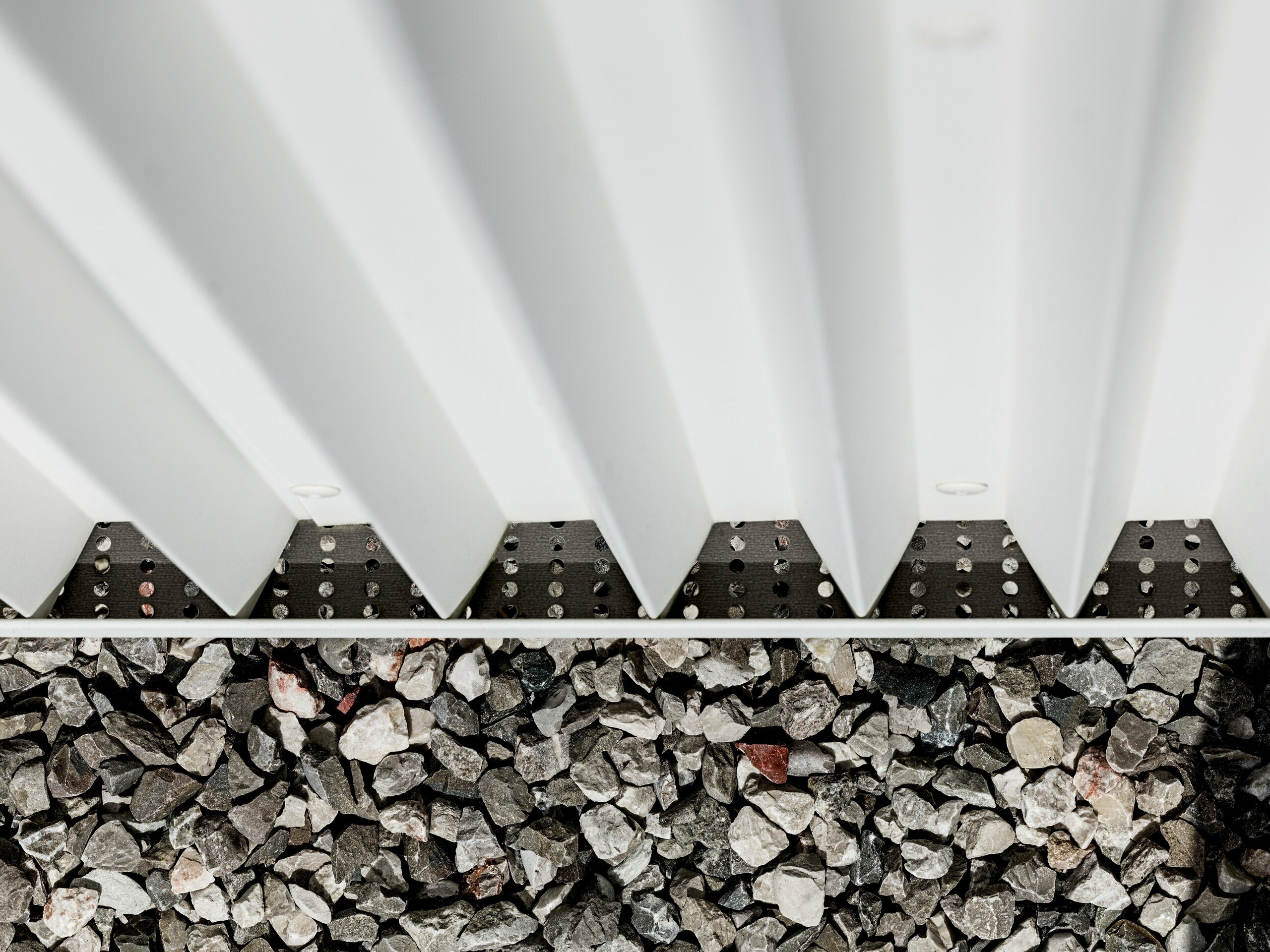 Obrázok zobrazuje detail vetranej fasády PREFALZ z čisto bieleho hliníka, ktorá je opatrená presne zloženými cikcakovými tvarmi. Táto inovatívna technika skladania zaručuje nielen opticky príťažlivú štruktúru, ale aj vynikajúce vetranie a odolnosť fasády voči poveternostným vplyvom. Spojenie fasádnych prvkov je realizované pomocou špeciálnych neviditeľných upevnení, ktoré umožňujú mimoriadne čistý a trvanlivý povrch. Spodná časť obrázku ukazuje štrkové pokrytie, ktoré slúži na odvodnenie a stabilizáciu okolia budovy, čo ďalej podčiarkuje funkčnosť dizajnu fasády. Použitím PREFALZ sa rovnako spĺňajú estetické nároky aj technické požiadavky, čo robí tento materiál ideálnou voľbou pre modernú a udržateľnú architektúru.