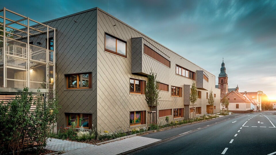 V jemnom svetle zapadajúceho slnka sa detské jasle v Alzenau ukazujú zo svojej najkrajšej stránky. Fasáda z bronzových PREFA šablón 44 × 44 odráža večerné svetlo a zdôrazňuje vysokokvalitný, štruktúrovaný povrch modernej hybridnej budovy. Jasne členitá architektúra s posunutými okennými pásmi a vertikálnymi drevenými lamelami vytvára zaujímavý kontrast s historickým kostolom v pozadí. Vľavo na obrázku zabezpečuje filigránová kovová konštrukcia ľahkú priehľadnosť vonkajšieho schodiska. Upravený zelený pás pozdĺž budovy podčiarkuje ekologický charakter. Tento obrázok pôsobivo spája modernú stavebnú kultúru, udržateľnú technológiu PREFA a harmonické začlenenie do historického obrazu mesta Alzenau.