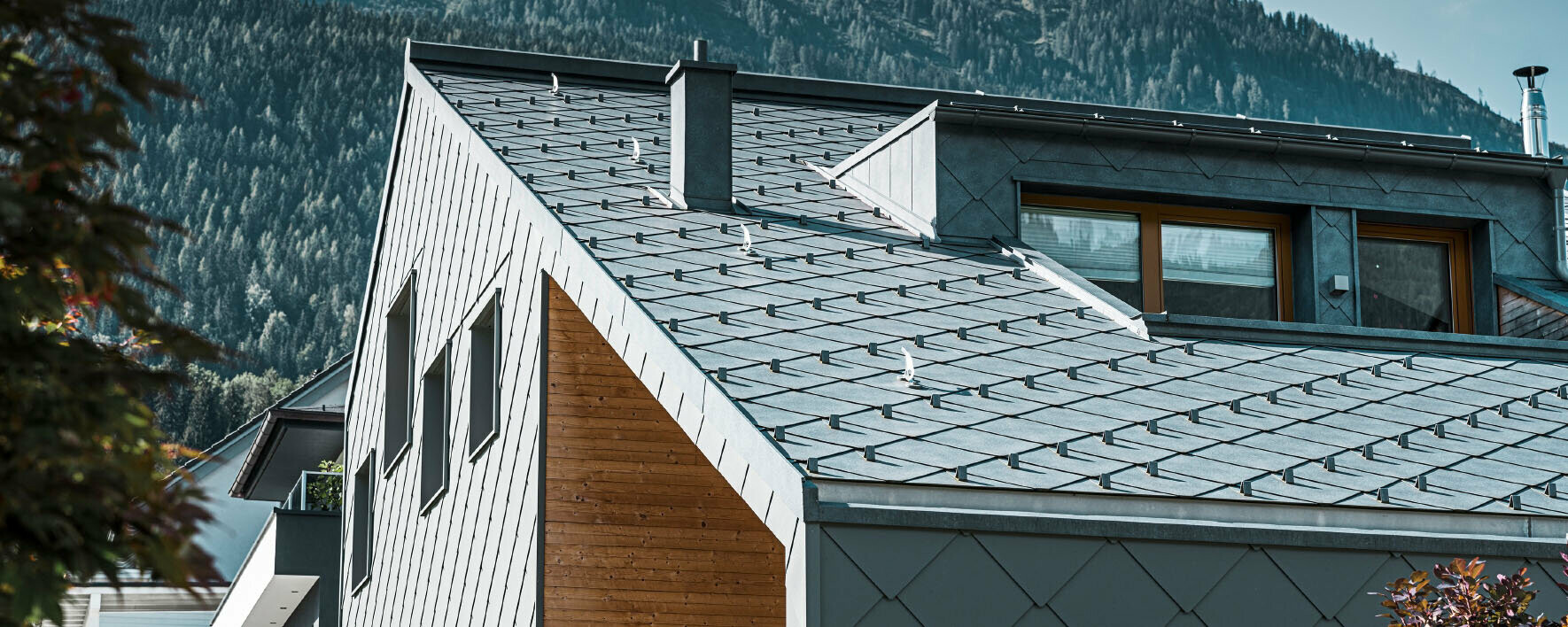 Dachansicht eines modernen Hauses mit hochwertigem PREFA Dachmaterial in schiefergrauer Optik. Das präzise verlegte, langlebige Stehfalzsystem zeigt die exzellente Handwerkskunst im Bereich Spenglerarbeiten und Dachdeckungen, ideal für anspruchsvolle Bauprojekte. Die natürliche Umgebung im Hintergrund betont die harmonische Integration des Daches in die Architektur.