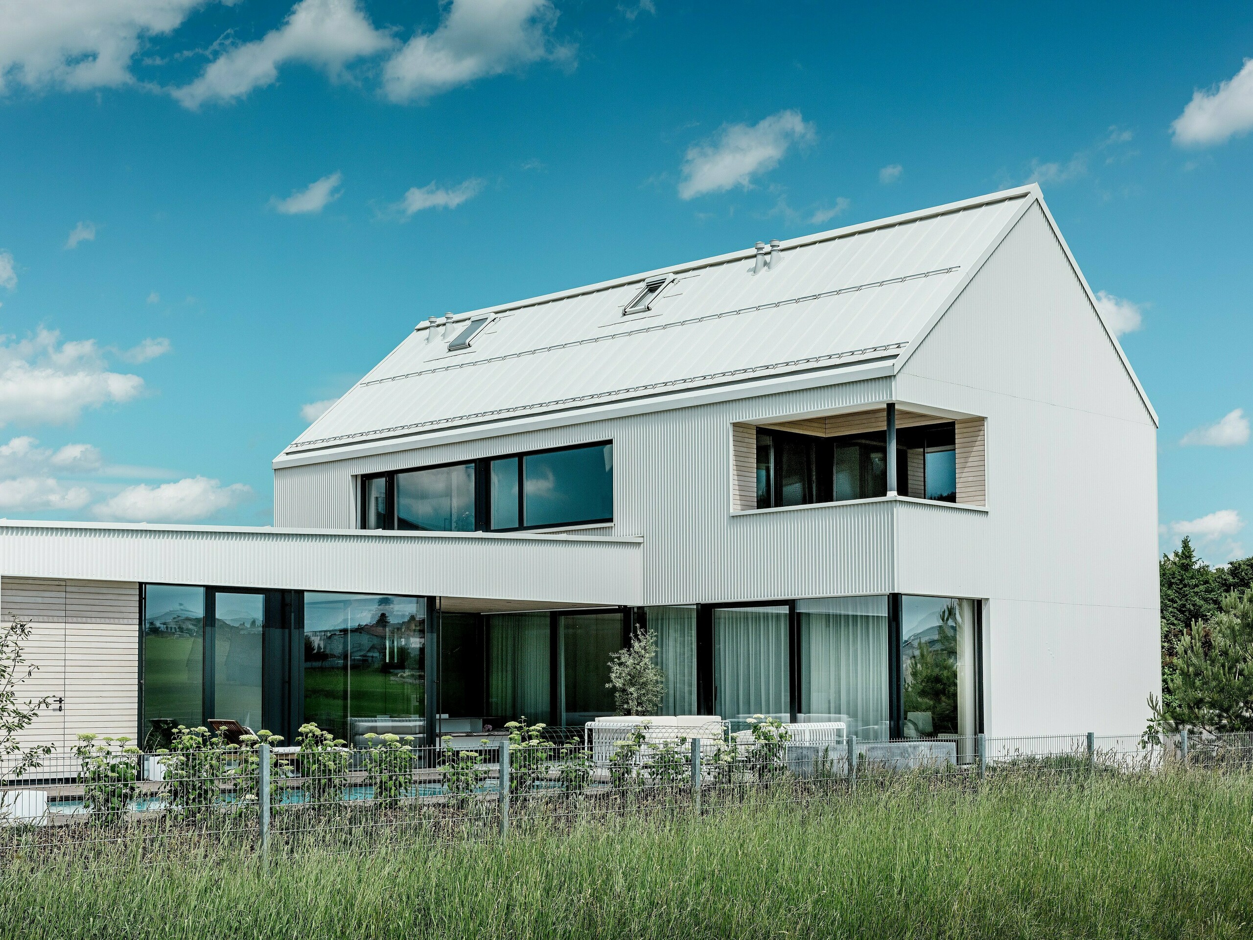 Tento impozantný rodinný dom vo Freistadte zaujme svojou elegantnou architektúrou, pri ktorej bol na fasádu a strechu použitý čisto biely hliník PREFALZ. Fasáda so zigzagovým skladaním vytvára moderný geometrický vzhľad, ktorý harmonicky zapadá do prírodného prostredia. Veľké okná nielenže vpustia do interiéru veľa denného svetla, ale ponúkajú aj široký výhľad na okolitú krajinu. Strmá sedlová strecha a moderný balkón s dreveným obložením podčiarkujú minimalistický dizajn a udržateľnú konštrukciu domu. PREFALZ zaisťuje optickú stálosť a odolnosť voči poveternostným vplyvom a garantuje dlhú životnosť pri minimálnej údržbe.