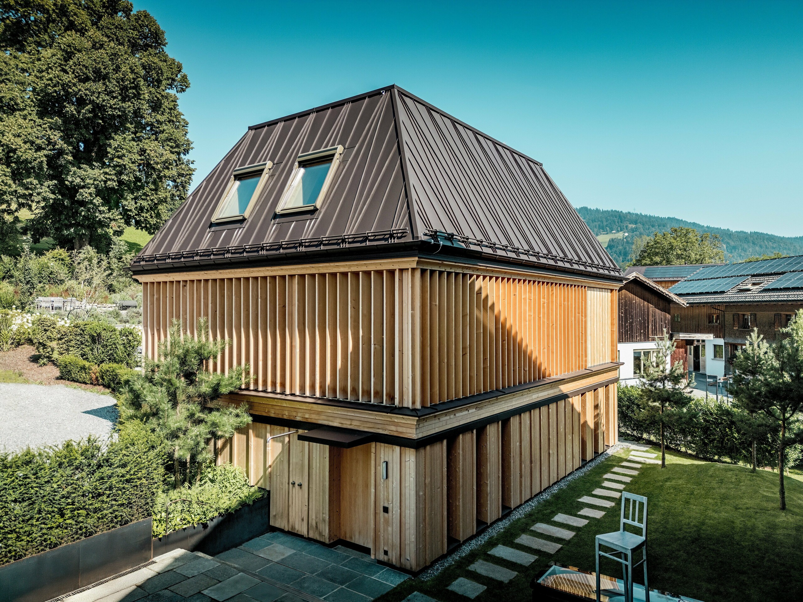 „Kúpeľný dom“ hotela Hirschen v Schwarzenbergu v jasnom, prirodzenom svetle. V centre pozornosti je výnimočná strecha PREFA so stojatými falcami v odtieni P.10 orieškovohnedá s presným prevedením a výraznými, čiastočne diagonálnymi stojatými falcami. Dve strešné okná zabezpečujú prirodzené osvetlenie interiéru. Fasáda je navrhnutá s vertikálne usporiadanými drevenými lamelami po celej výške poschodia, ktoré vynikajú ako po dizajnovej, tak aj po funkčnej stránke. V popredí vedú prírodné kamenné dosky cez upravenú záhradu. Obklopená tradičnými drevenými domami a miernymi kopcami sa táto moderná budova harmonicky začleňuje do obrazu Bregenzerwaldu.