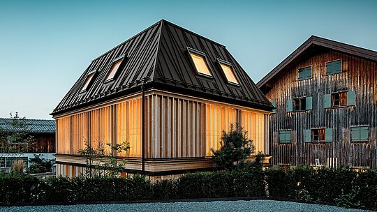 Ocenená „kúpeľná budova“ hotela Hirschen v Schwarzenbergu v atmosférickom večernom svetle. V popredí dominuje moderná valbová strecha s výrazným PREFA stojatým falcom v odtieni P.10 orieškovohnedá – skutočné architektonické vyhlásenie, ktoré zaujme svojimi nekonvenčnými líniami a sklonom 60 stupňov. Fasáda z vertikálnych drevených lamiel po celej výške poschodia harmonicky dopĺňa historický komplex a spája moderný minimalizmus s tradíciou staviteľstva v regióne Bregenzerwald. V pozadí je vidieť pamiatkovo chránenú hlavnú budovu – silný kontrast starého a nového. Úspešný príklad budovania orientovaného na budúcnosť s použitím vysoko kvalitných materiálov PREFA.