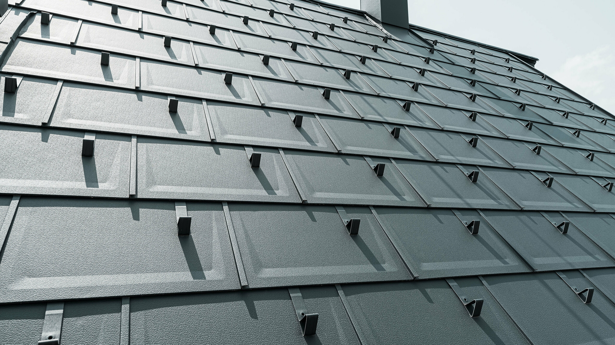 Detailný pohľad na strešný panel PREFA R.16 v farbe P.10 antracit. Na streche sú viditeľné snehové zábrany PREFA a komín.