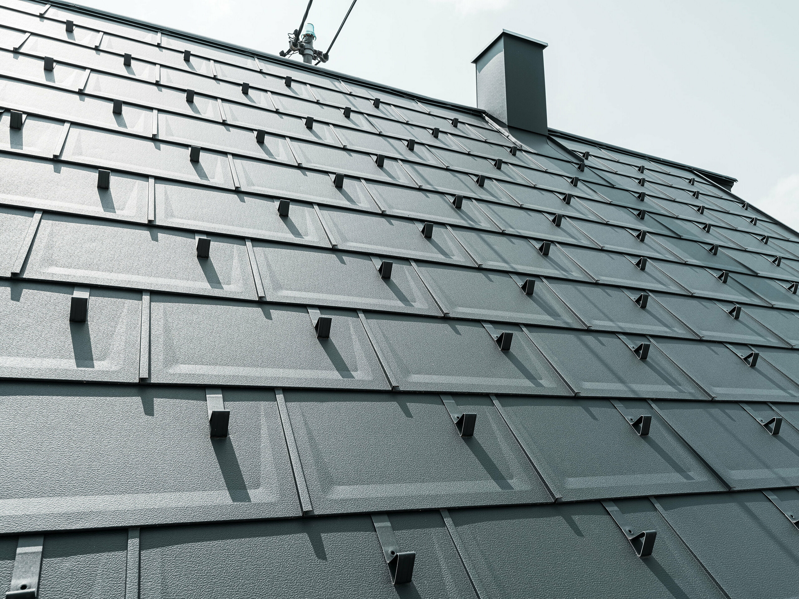 Detailný pohľad na strešný panel PREFA R.16 v farbe P.10 antracit. Na streche sú viditeľné snehové zábrany PREFA a komín.