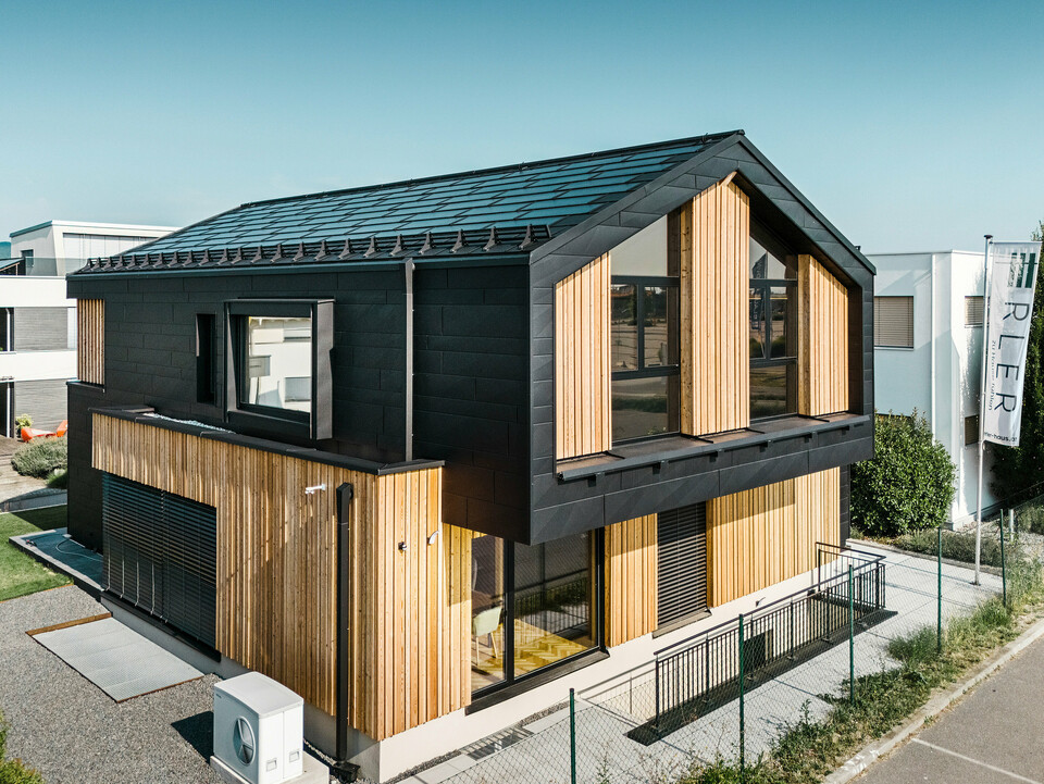 Predná časť modelového domu Autark v centre montovaných domov Blaue Lagune, Vösendorf (AT). Strecha je pokrytá integrovanými solárnymi panelmi PREFA a strešnými panelmi R.16 s kvalitným náterom P.10. Fasádu tvorí kombinácia vertikálnych drevených prvkov a PREFA Siding.X vo farbe P.10 čierna, ktorá dáva budove moderný a udržateľný charakter. Vlajka Erler Bau GmbH veje na pravom okraji obrázku.