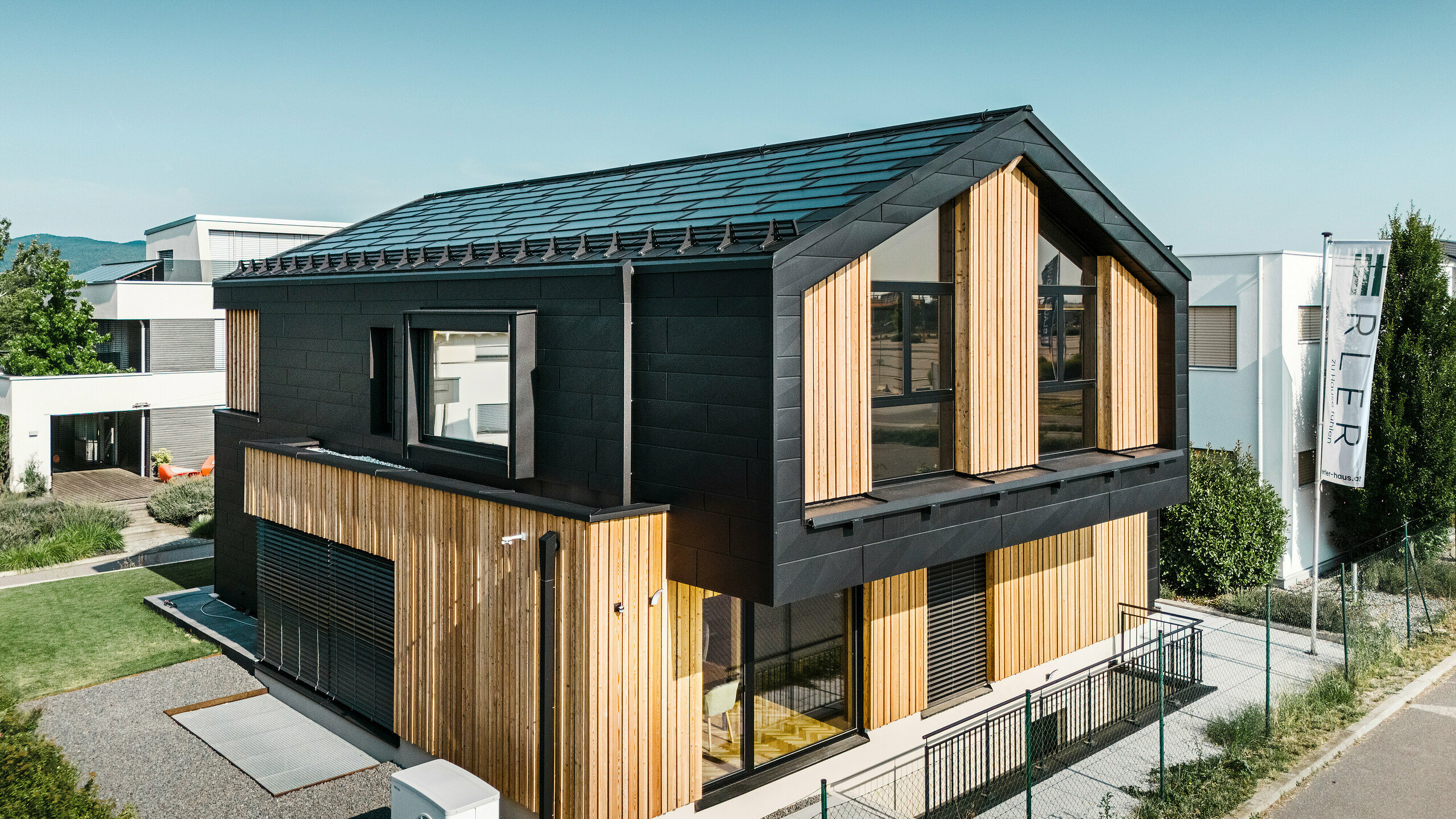 Predná časť modelového domu Autark v centre montovaných domov Blaue Lagune, Vösendorf (AT). Strecha je pokrytá integrovanými solárnymi panelmi PREFA a strešnými panelmi R.16 s kvalitným náterom P.10. Fasádu tvorí kombinácia vertikálnych drevených prvkov a PREFA Siding.X vo farbe P.10 čierna, ktorá dáva budove moderný a udržateľný charakter. Vlajka Erler Bau GmbH veje na pravom okraji obrázku.