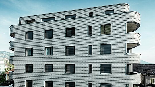 Pohľad z prednej strany domu Bernerhaus v Buchsi s jemne zaoblenými rohmi budovy a súvislou fasádou z PREFA šindľov v odtieni P.10 svetlošedá. Hliníková fasáda položená vo forme šupín vytvára živý vzor a zdôrazňuje horizontálne línie zakrivených balkónových pásov. Presne integrované okenné otvory sa harmonicky začleňujú do homogénneho povrchu. Vďaka ľahkému, odolnému voči poveternostným vplyvom a trvácnemu hliníku PREFA vzniká obvodový plášť budovy s nízkymi nárokmi na údržbu, tvarovo stabilný a s výrazným vzhľadom v alpskom prostredí.