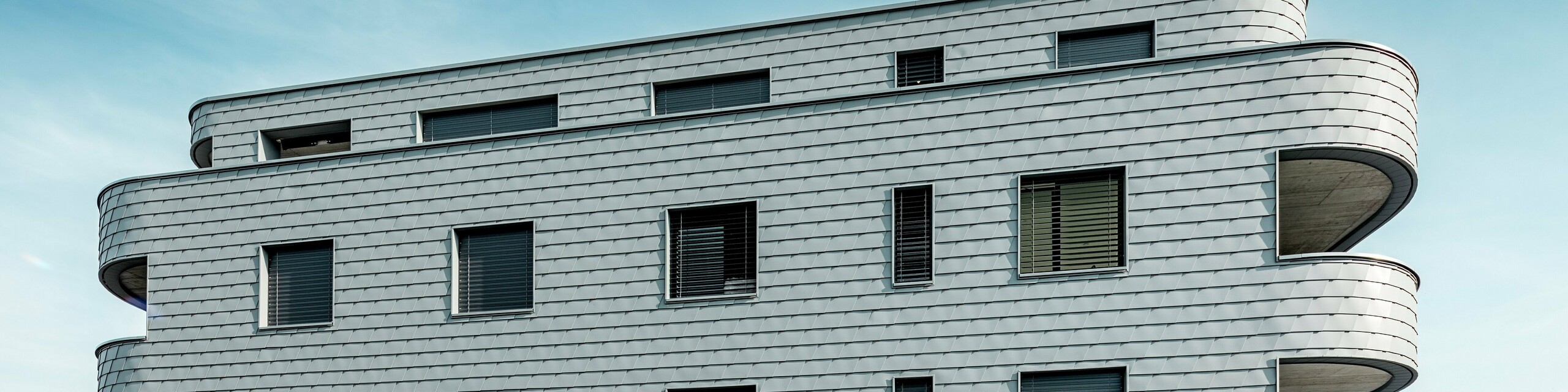 Pohľad z prednej strany domu Bernerhaus v Buchsi s jemne zaoblenými rohmi budovy a súvislou fasádou z PREFA šindľov v odtieni P.10 svetlošedá. Hliníková fasáda položená vo forme šupín vytvára živý vzor a zdôrazňuje horizontálne línie zakrivených balkónových pásov. Presne integrované okenné otvory sa harmonicky začleňujú do homogénneho povrchu. Vďaka ľahkému, odolnému voči poveternostným vplyvom a trvácnemu hliníku PREFA vzniká obvodový plášť budovy s nízkymi nárokmi na údržbu, tvarovo stabilný a s výrazným vzhľadom v alpskom prostredí.