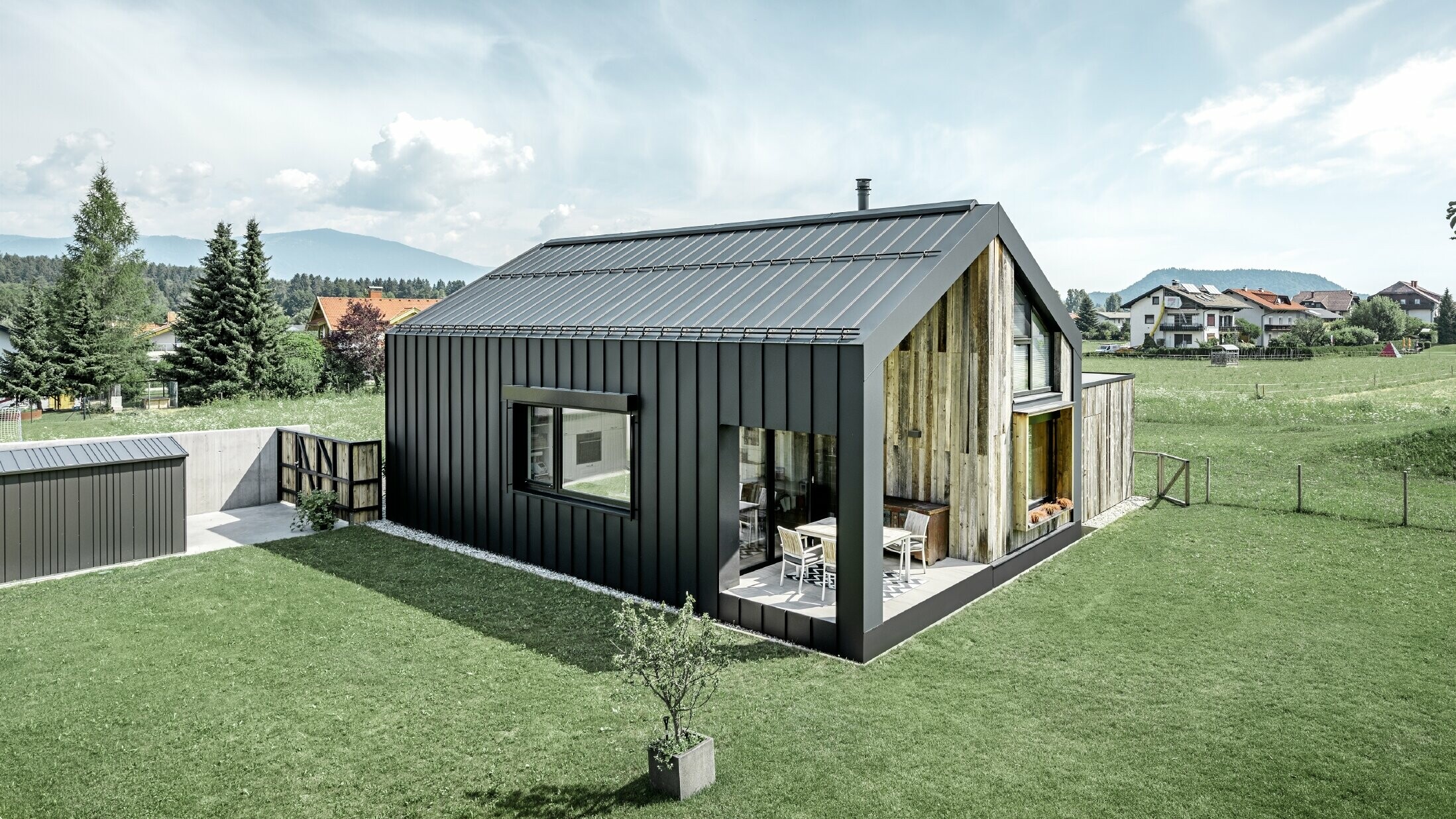 Rodinný dom so sedlovou strechou, strecha a fasáda sú pokryté so systémom PREFALZ od firmy PREFA vo farebnom vyhotovení P.10 antracitová. Pásy plochy strechy sa ťahajú tiež ďalej cez fasádu. Časť fasády je obložená so zvetraným drevom.