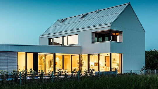 Obrázok ukazuje moderný rodinný dom vo Freistadte z perspektívy, ktorá zdôrazňuje harmonické spojenie architektúry a prírody. Výrazná fasáda PREFALZ v čistej bielej farbe okamžite upúta pozornosť, zatiaľ čo veľkorysé sklenené plochy dodávajú interiéru otvorený a príjemný vzhľad. Strmá sedlová strecha, tiež pokrytá hliníkom PREFALZ, zaisťuje dynamický záver a chráni budovu pred vplyvmi počasia. Štýlový minimalistický dizajn je zdôraznený teplými drevenými prvkami na balkóne a terase, čo vytvára príjemný kontrast. Budova je nielen esteticky príťažlivá, ale aj funkčná a udržateľná vďaka odolným a ľahko udržiavateľným hliníkovým produktom PREFALZ.