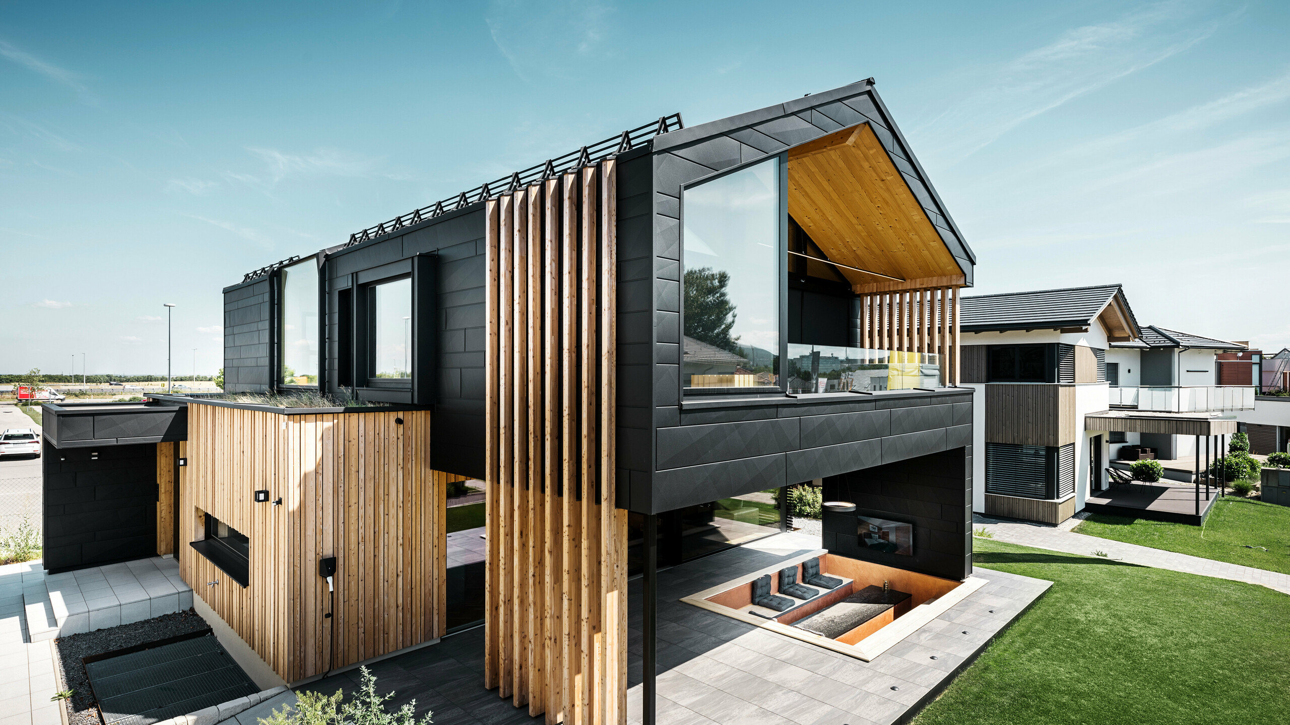 Bočný pohľad na výstavný dom Autark v Modrej lagúne v rakúskom Vösendorfe. Horné poschodie zaujme veľkými oknami a modernou hliníkovou fasádou z panelov PREFA Siding.X v čiernej farbe P.10 v kombinácii s vertikálnymi drevenými prvkami. Priestranná terasa na prízemí so zapusteným sedením plynulo zapadá do architektúry. Štýlový a udržateľný dizajn domu pôsobivo kombinuje hliník a drevo.