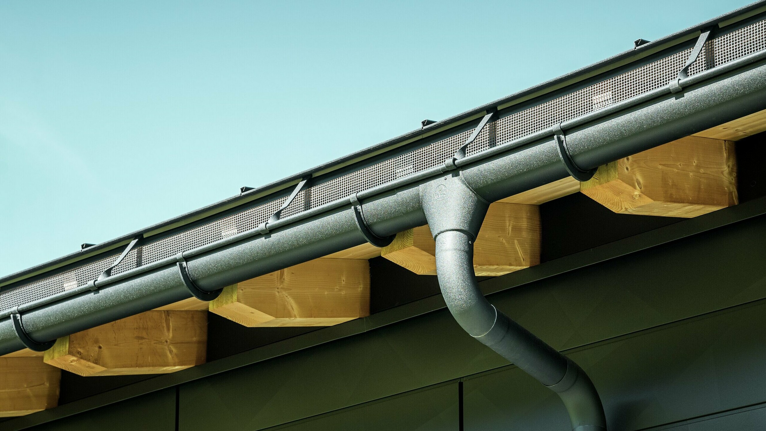 Detailný pohľad na fasádu moderného rodinného domu v Ascharine vo Švajčiarsku. Bočné steny budovy boli obložené výraznými fasádnymi panelmi PREFA Siding.X v čiernej farbe P.10. Farebne zladený je aj odvodňovací systém PREFA so žľabom a zvodom.