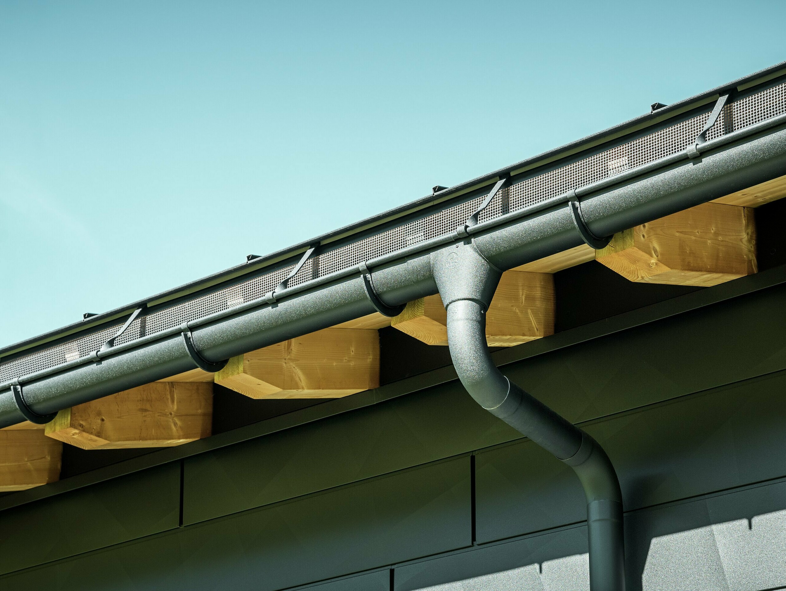 Detailný pohľad na fasádu moderného rodinného domu v Ascharine vo Švajčiarsku. Bočné steny budovy boli obložené výraznými fasádnymi panelmi PREFA Siding.X v čiernej farbe P.10. Farebne zladený je aj odvodňovací systém PREFA so žľabom a zvodom.