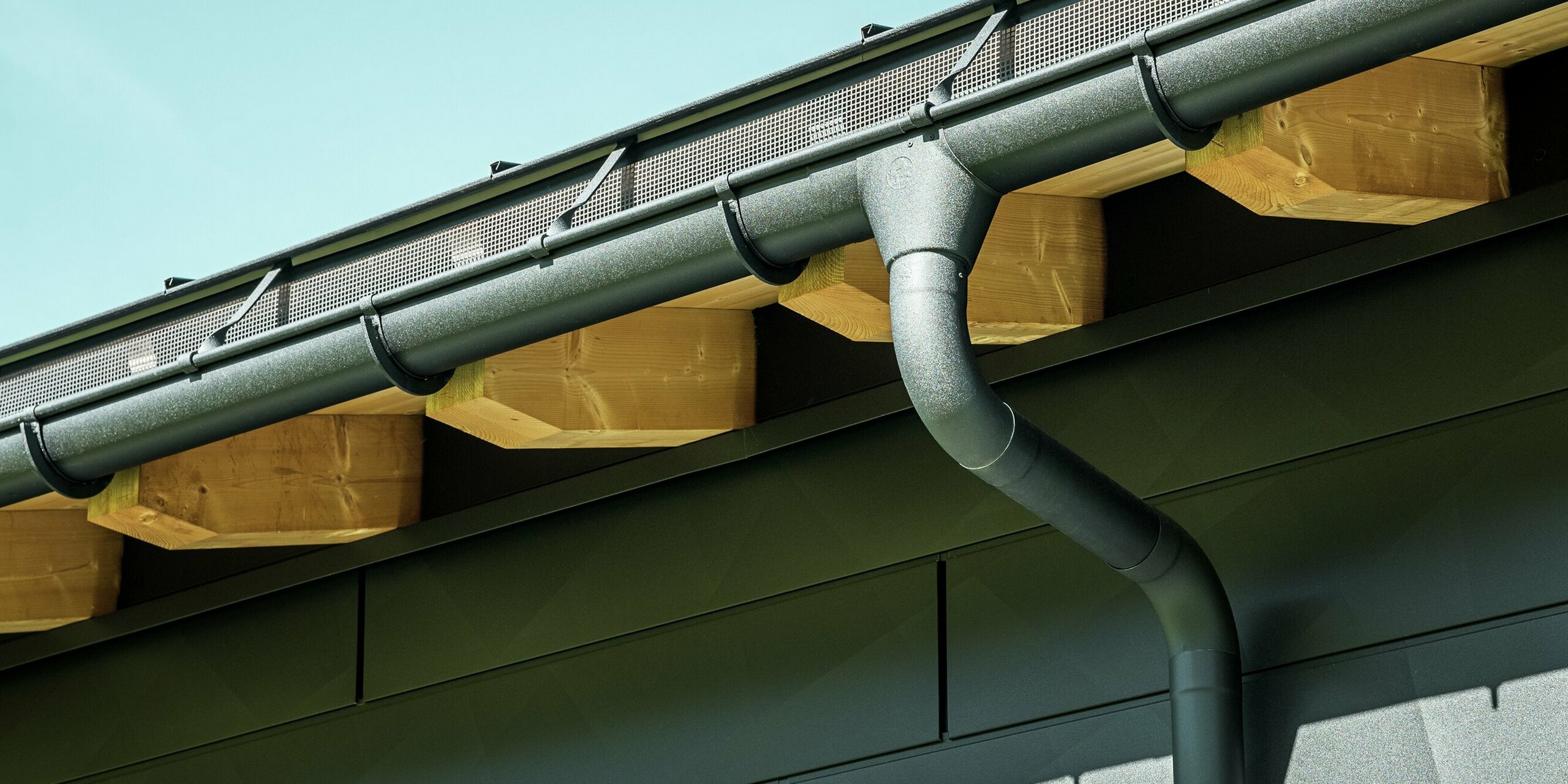 Detailný pohľad na fasádu moderného rodinného domu v Ascharine vo Švajčiarsku. Bočné steny budovy boli obložené výraznými fasádnymi panelmi PREFA Siding.X v čiernej farbe P.10. Farebne zladený je aj odvodňovací systém PREFA so žľabom a zvodom.