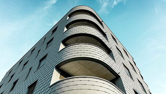 Dynamický pohľad na spodnú časť modernej obytnej a obchodnej budovy s výrazne zaobleným rohom a plynulými balkónovými pásmi. Šupinová fasáda z PREFA fasádnych šindľov v odtieni P.10 svetlošedá pokrýva budovu ako precízne spracovaná hliníková koža a zdôrazňuje jej jemné polomery. Homogénny povrch podčiarkuje čisté línie, zatiaľ čo ľahký, odolný voči poveternostným vplyvom hliník PREFA zaručuje aj pri silných zaobleniach najvyššiu tvarovú stabilitu, dlhú životnosť a nízke nároky na údržbu – architektonický pohľad upútavajúci pred žiarivo modrou oblohou.