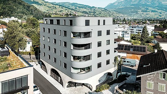 Letecký pohľad na Bernerhaus v Buchsi s výraznou trojuholníkovou formou budovy a jemne zaoblenými balkónmi uprostred alpského krajiny. Súvislá fasáda z PREFA fasádnych šindľov v odtieni P.10 svetlošedá obklopuje budovu ako šupinatá hliníková koža a vytvára živú hru svetla a štruktúry. Nad masívnym betónovým podstavcom s arkádami pôsobí ľahká, odolná proti poveternostným vplyvom hliníková fasáda PREFA mimoriadne precízne, trvalo a s nízkymi nárokmi na údržbu – moderný pohľadový prvok v mestskom prostredí.