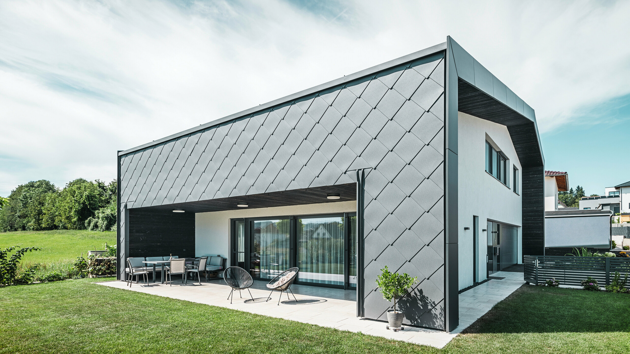 Udržiavaná záhradná časť rodinného domu v Aschachu. Objekt je štýlovo obložený tmavosivými hliníkovými fasádnymi šablónami PREFA 44 × 44. Krytá terasa ponúka útulné posedenie s výhľadom na svieži zelený trávnik. Elegantný hliníkový obklad v kombinácii s bielymi omietnutými plochami zabezpečuje modernú architektúru odolnú voči poveternostným vplyvom. Veľké okná prepúšťajú do interiéru dostatok denného svetla a vytvárajú otvorenú, svetlom zaliatu atmosféru.