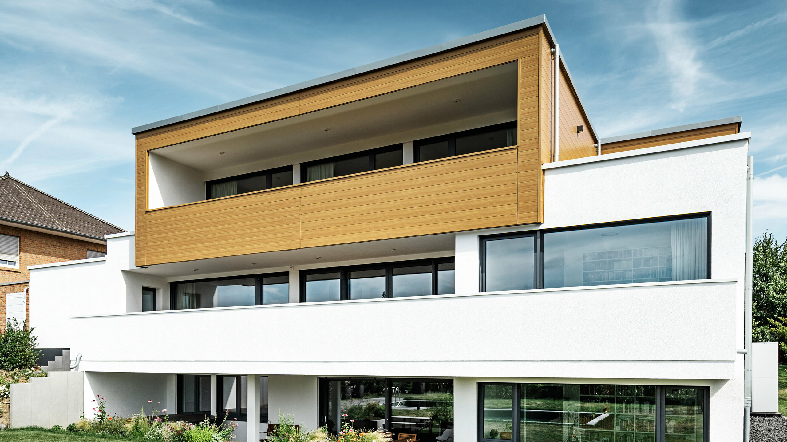 Zadná časť moderného rodinného domu v Usingene, Nemecko. V dolnej časti obrázka je vidieť zelený trávnik, bazén a mladý záhradný strom. Pútavý dom má biely základ budovy z omietnutej fasády, hliníkovú fasádu na hornom poschodí z PREFA Sidings v dub prírodný farbe a veľké okenné plochy s rámom v antracitovej farbe. Z diaľky je na ľavom okraji obrázka vidieť susedný dom s tehlovou fasádou.