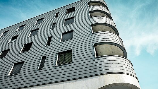 Detailný záber na Bernerhaus v Buchsi s výrazne zaobleným rohom budovy a plynulými otvormi balkónov. Fasáda z PREFA šindľov v odtieni P.10 svetlošedá pokrýva jasne štruktúrovanú kubatúru ako precízne spracovaná hliníková koža. Šupinatý povrch zdôrazňuje jemné polomery a vytvára živú hru svetla a tieňa. Vďaka ľahkému, odolnému voči poveternostným vplyvom a trvácnemu hliníku PREFA vzniká homogénny plášť budovy s nízkymi nárokmi na údržbu – technicky náročný a architektonicky skutočne pútavý.