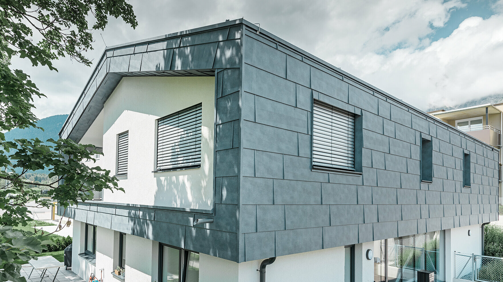 Moderný rodinný dom bez previsnutej strechy; horné poschodie bolo opláštené fasádnymi panelmi FX.12 od firmy PREFA vo farbe kamenná šedá, zvyšok fasády bol omietnutý nabielo.