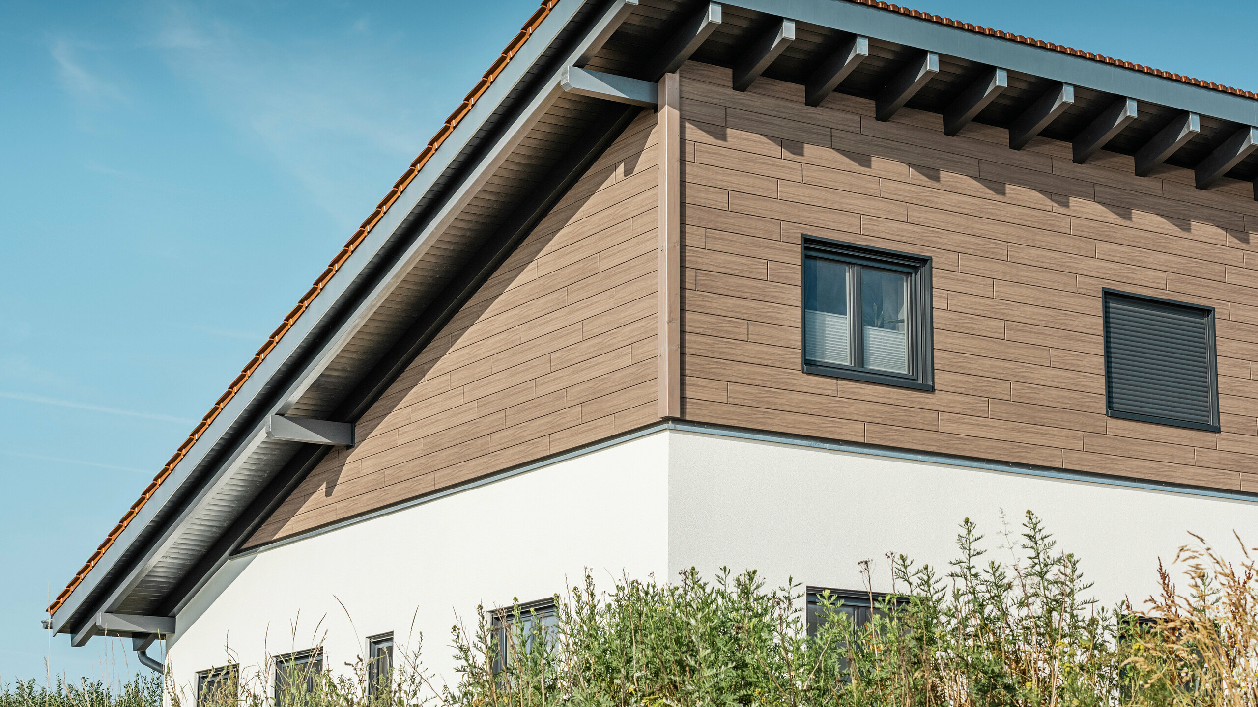 Schräger Blick von unten auf das Obergeschoss eines Einfamilienhauses in Bruck. Das obere Stockwerk ist mit PREFA Sidings in einer Holzoptik umhüllt. Zu sehen ist das Pultdach, das auf der Gebäuderückseite nach unten verläuft.