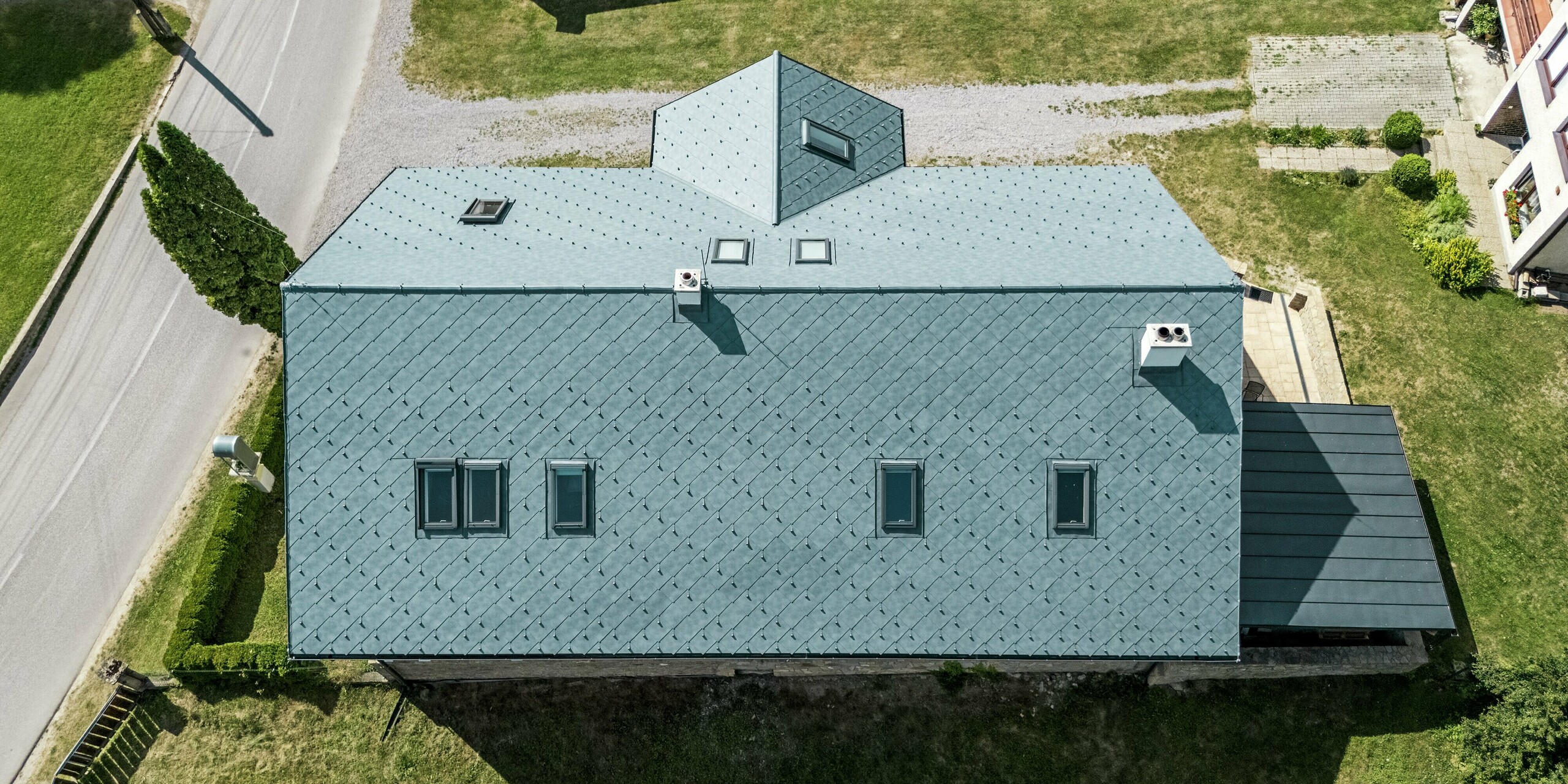 Das Bild zeigt die beeindruckende Ansicht des Daches des Handwerkerhauses in Klin, Slowakei, aus der Vogelperspektive. Die symmetrische Anordnung der PREFA Dachrauten 44 × 44 in P.10 Steingrau unterstreicht die elegante Architektur des Hauses. Die zahlreichen Dachfenster fügen sich harmonisch in die Dachstruktur ein und sorgen für eine helle, freundliche Atmosphäre im Inneren. Das Aluminiumdach bietet neben einer ästhetischen Aufwertung auch praktischen Nutzen: Es ist besonders langlebig, korrosionsbeständig und leicht, was es zur idealen Wahl für die Sanierung eines historischen Gebäudes macht. Diese Dachlösung verbindet perfekte Optik mit optimaler Funktionalität und Widerstandsfähigkeit gegenüber Witterungseinflüssen.