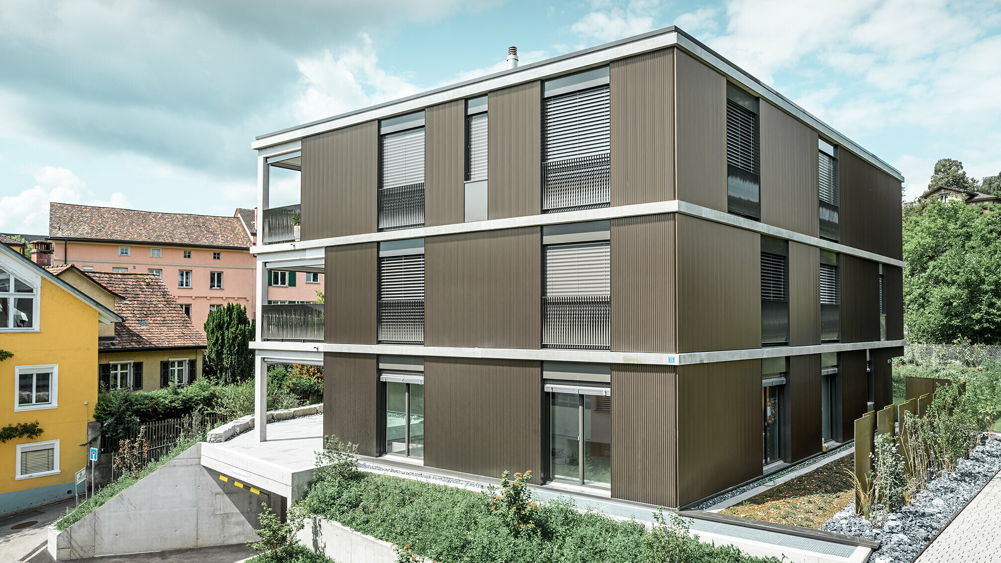 Výrazná hliníková fasáda tohto viacgeneračného domu v Schaffhausene je vybavená elegantným prelamovaným profilom PREFA Zackenprofil v farbe Colinal. Vďaka vertikálnemu usporiadaniu extrudovaných hliníkových profilov pôsobí budova opticky kompaktnejšie a štruktúra fasády získava harmonickú rovnováhu. Elegantný dizajn sa vkusne začleňuje do okolia a dodáva budove modernú eleganciu, ktorá je nielen vizuálne príťažlivá, ale aj funkčná. Perspektíva ukazuje bytový dom z prednej strany s vjazdom do garáže. V pozadí sú viditeľné stromy, ďalšie budovy a veža kostola.