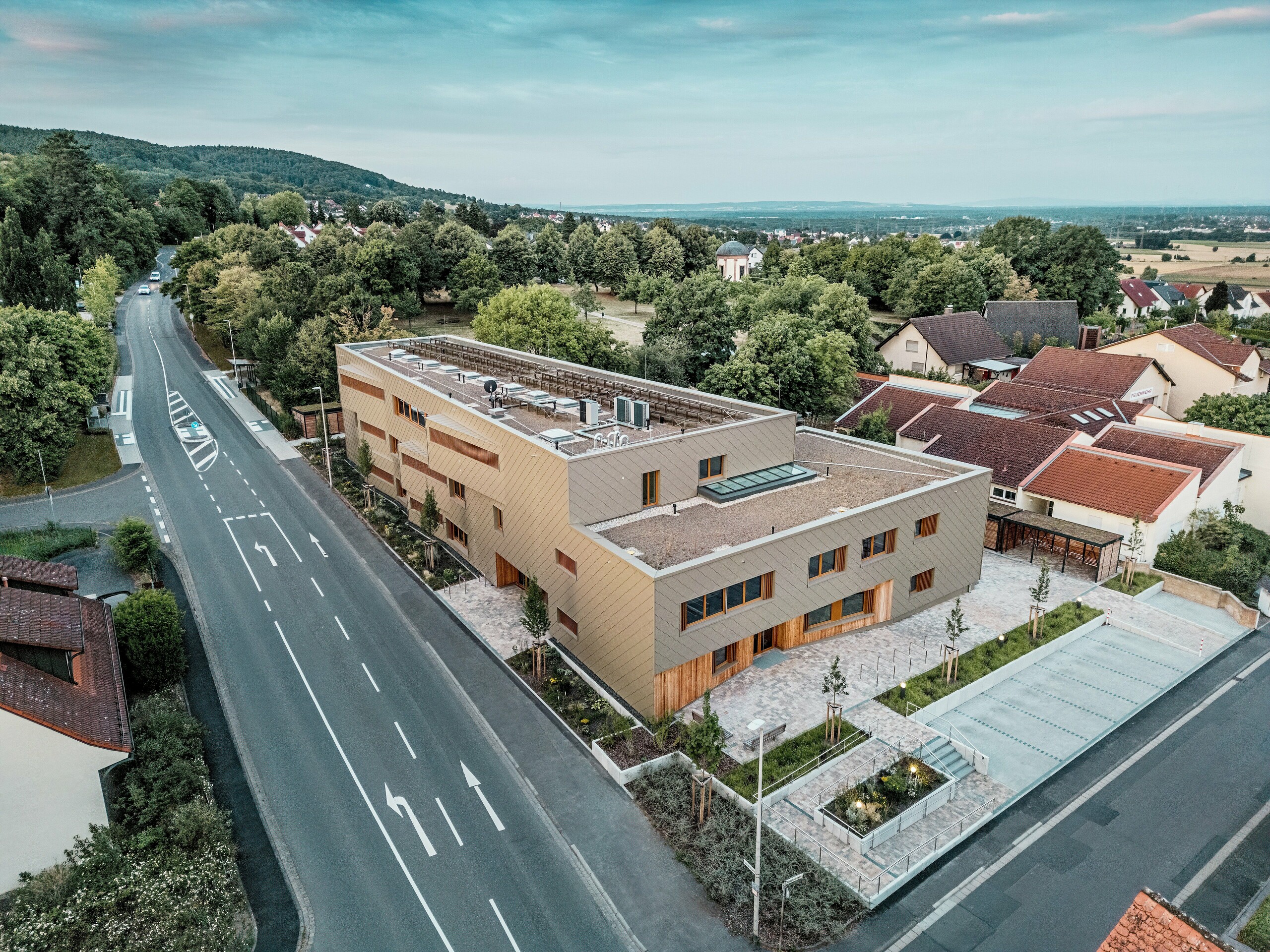 Impozantné celkové riešenie materskej školy v Alzenau je viditeľné z leteckého pohľadu. L tvarová hybridná budova je harmonicky zasadená do pokojnej obytného štvrti na okraji Spessartu. Fasáda z bronzovo sfarbených PREFA fasádnych šablón 44 × 44 dodáva stavbe výrazný a kvalitný vzhľad a zdôrazňuje modernú architektúru. Zelené strechy, priestranné vonkajšie plochy a jasné riešenie dopravy podčiarkujú udržateľný koncept celku. Kombinácia trvácneho hliníka, dreva a recyklovaného betónu spĺňa najvyššie ekologické štandardy. Táto perspektíva zvýrazňuje premyslenú urbanistickú integráciu a multifunkčný charakter budovy – perspektívny projekt pre vzdelávanie, bývanie a udržateľnosť.