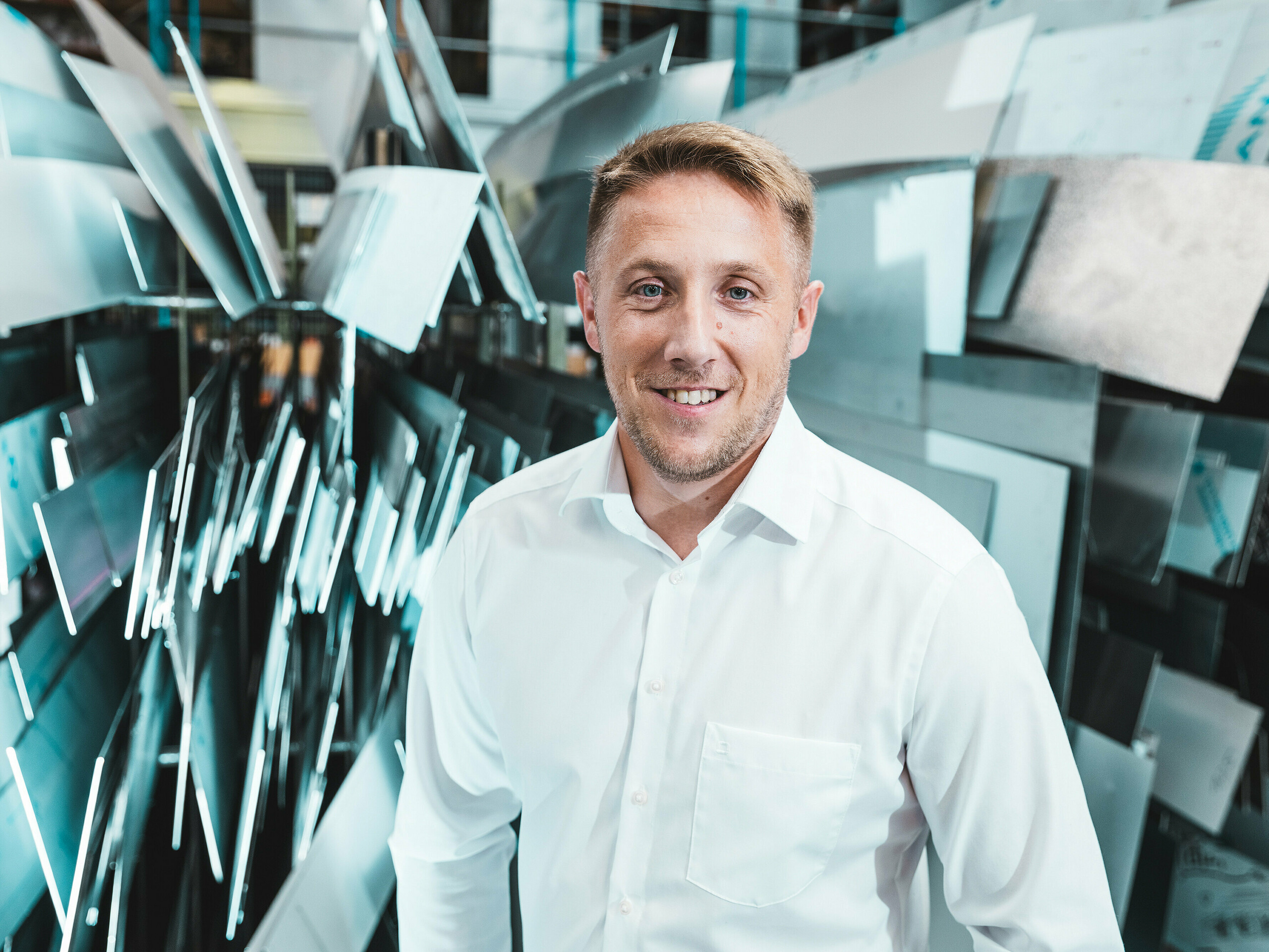 Sebavedomo a priateľsky stojí projektový vedúci Marcel Schmidt z klampiarskej firmy ASA Schüßler pred impozantnou stenou z rôzne tvarovaných hliníkových prvkov. Dynamicky usporiadané kovové platne efektne odrážajú svetlo a symbolizujú inovácie, precíznosť a kvalitu v spracovaní kovov. Muž v bielej košeli stelesňuje odbornosť a modernú výrobnícku excelentnosť. V pozadí sa ukazuje široká škála možností spracovania hliníka – od strešných a fasádnych systémov až po zákazkové riešenia na mieru.