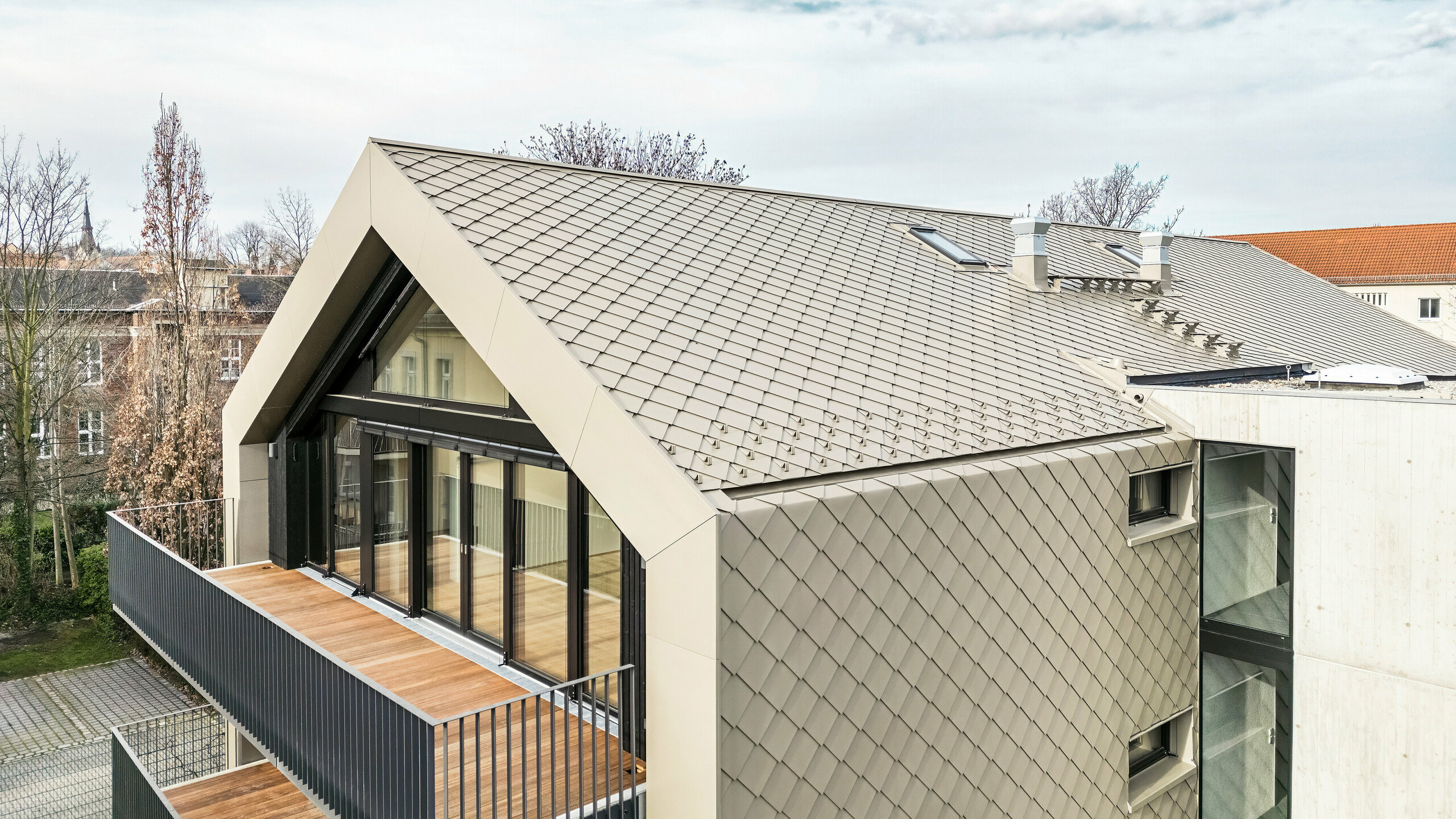 Pohľad z vtáčej perspektívy na moderný bytový dom na Chemnitzer Straße (Drážďany) s obvodovým plášťom budovy z hliníkových šablón PREFA 29 × 29 v bronzovej farbe. Na obrázku je vidieť elegantnú strešnú konštrukciu, veľké presklené priečelia a balkóny, ako aj harmonické začlenenie budovy do okolia. Vysokokvalitné materiály zdôrazňujú odolnú architektúru budovy.