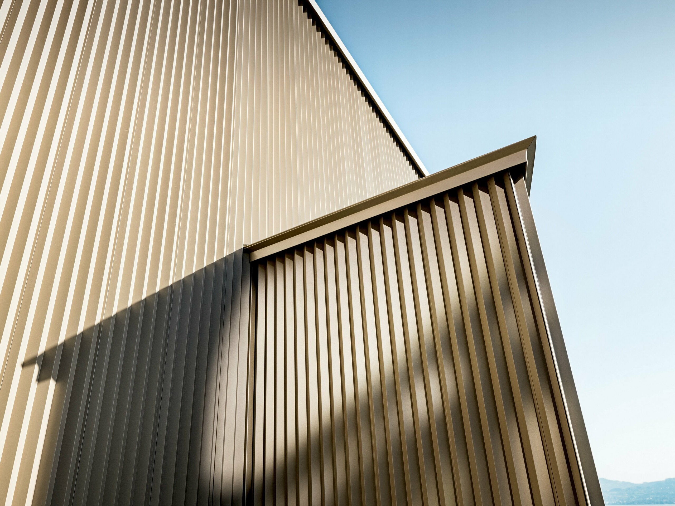 Elegantná hliníková fasáda PREFA v bronzovom vyhotovení na obytnom dome v St. Gingolph, navrhnutá architektkou Sophie Morard. Vertikálne prelamované profily vytvárajú fascinujúcu hru svetla, tieňa a štruktúry, ktorá dodáva fasáde hĺbku a dynamiku. Vysoko kvalitný povrch s práškovou farbou zaisťuje dlhodobú ochranu a štýlovú estetiku – ideálne pre náročné projekty v oblasti bývania. Bronzovo lesklý dizajn sa harmonicky začleňuje do alpského prostredia a robí z malého domu architektonický skvost s veľkým účinkom.