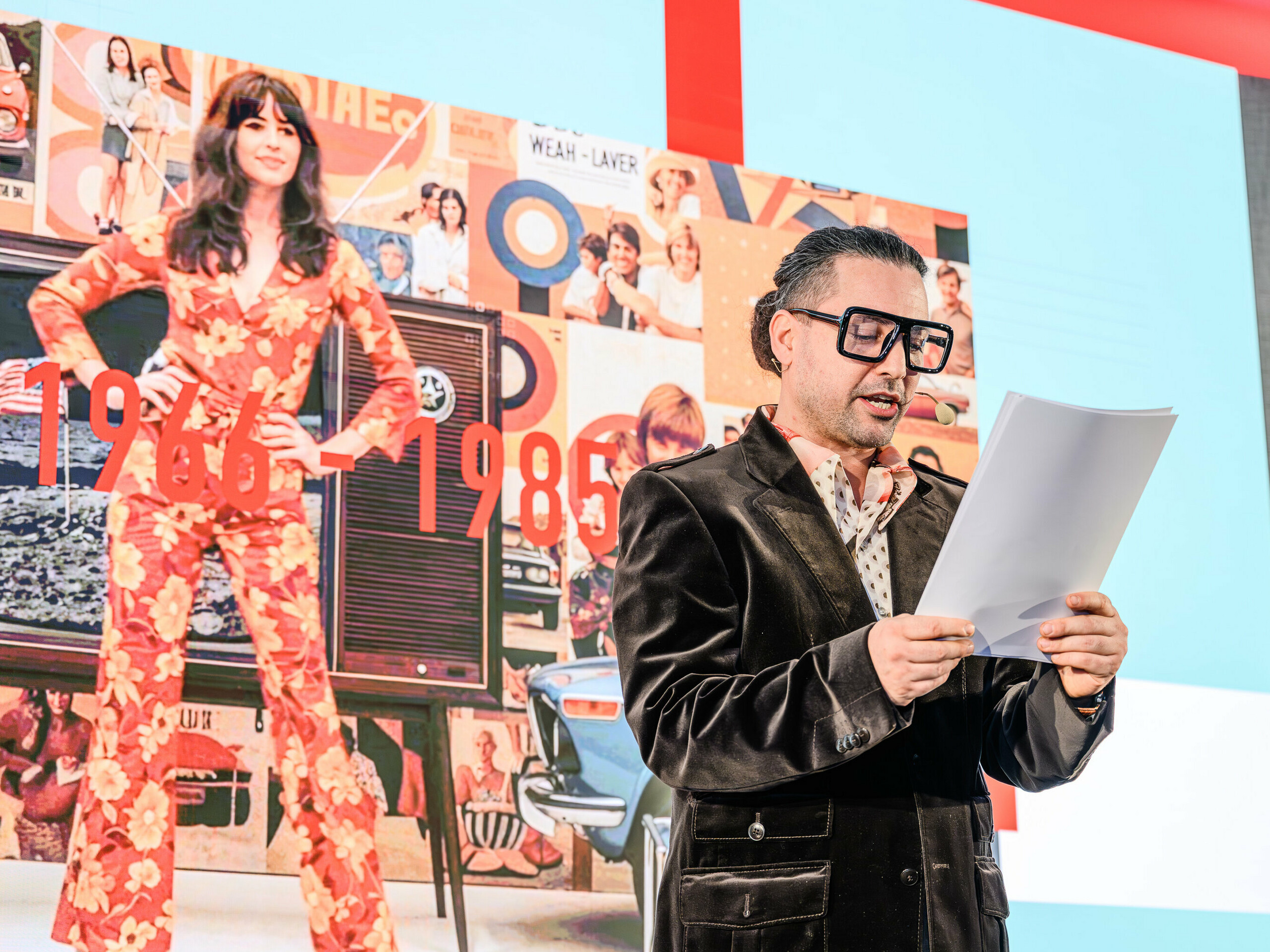 Cesta časom na pódiu prezentácie PREFARENZEN 2026: Charizmatický Serkan Temel v retro štýle sprevádza publikum cez desaťročia. Zamerané je na roky 1966 až 1985, ktoré sú znázornené na koláži v pozadí s ikonami dizajnu a spoločenskými trendmi tejto éry. Rovnako ako produkty PREFA, ktoré pretrvávajú trendy so štýlom a podstatou. Prezentácia ukazuje, ako sa architektúra včerajška stáva inšpiráciou pre riešenia zajtrajška – s novým výkladom hliníka.