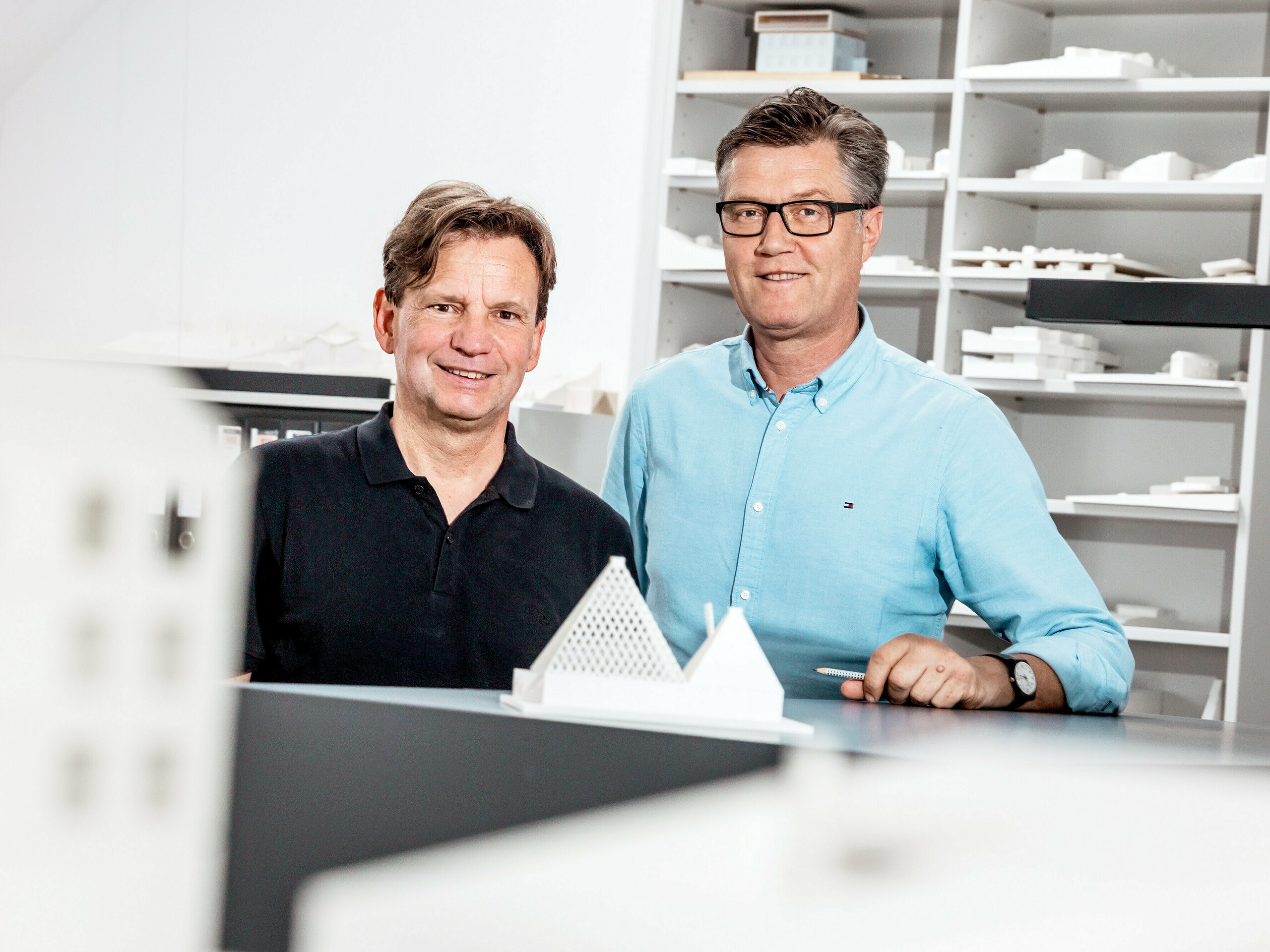 Na fotografii sú architekti zo štúdia Schneider Lengauer Pühringer, ktorí hrdo pózujú vo svojej kancelárii pred zbierkou architektonických modelov. Navrhli moderný rodinný dom vo Freistadte s názvom projektu „One Shade Of White” a sú známi svojím inovatívnym a estetickým prístupom k architektúre. Modely v pozadí odrážajú ich kreatívne koncepty a demonštrujú ich schopnosť kombinovať moderné dizajnové nápady s funkčnými riešeniami. Obaja sú oblečení v ležérnom štýle, čo podčiarkuje uvoľnenú, ale zároveň profesionálnu atmosféru v ich pracovnom prostredí.