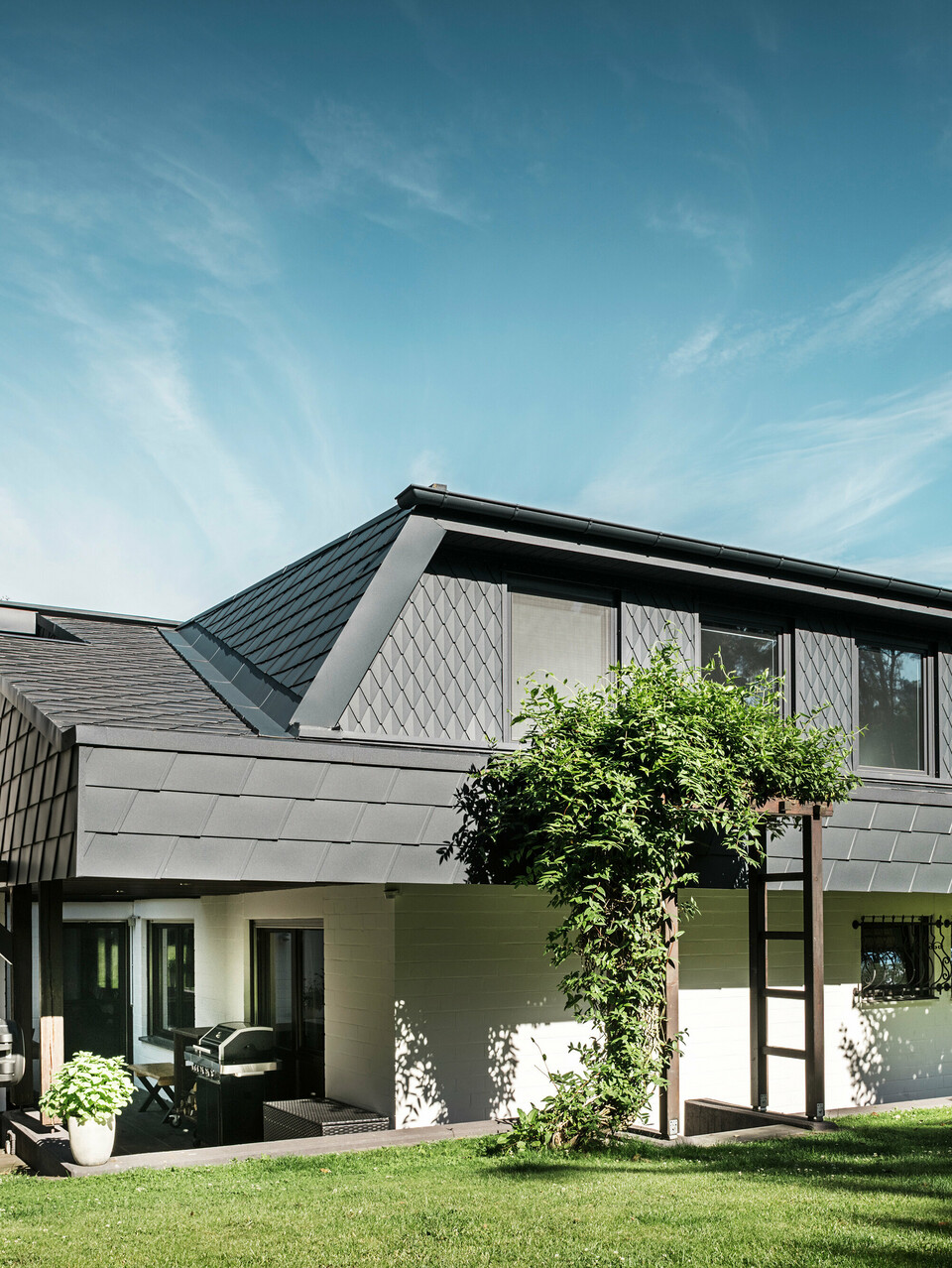 Bočný pohľad na rodinný dom v Mechernich so strešnými šindľami PREFA DS.19 a malými strešnými šablónami v antracitovej farbe P.10. Trapézové vikiere sú opláštené strešnými šablónami z robustného plechu, zatiaľ čo strecha je pokrytá šindľom. Modernú architektúru odolnú voči poveternostným vplyvom dopĺňa zelená pergola a stromy v záhradnej časti.