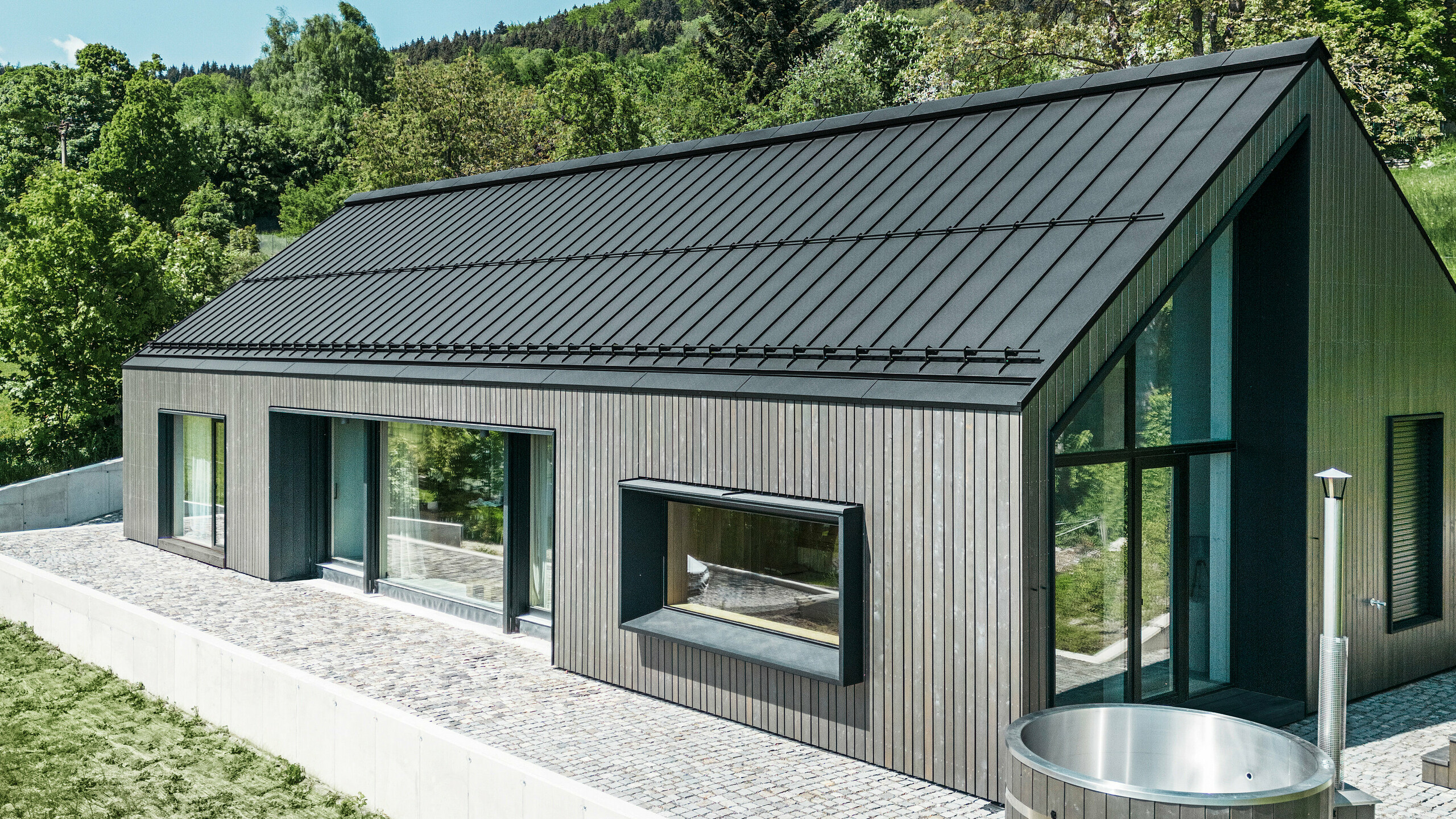 Ein modernes Holzmassivhaus in Kuncice (CZ) wurde mit großen Fensterflächen ausgestattet. Das dunkle Holzmassivhaus verfügt über ein Dachsystem aus PREFALZ in der Farbe P. 10 Schwarz. Im hinteren Außenbereich steht ein Whirlpool mit einer Holzverkleidung.