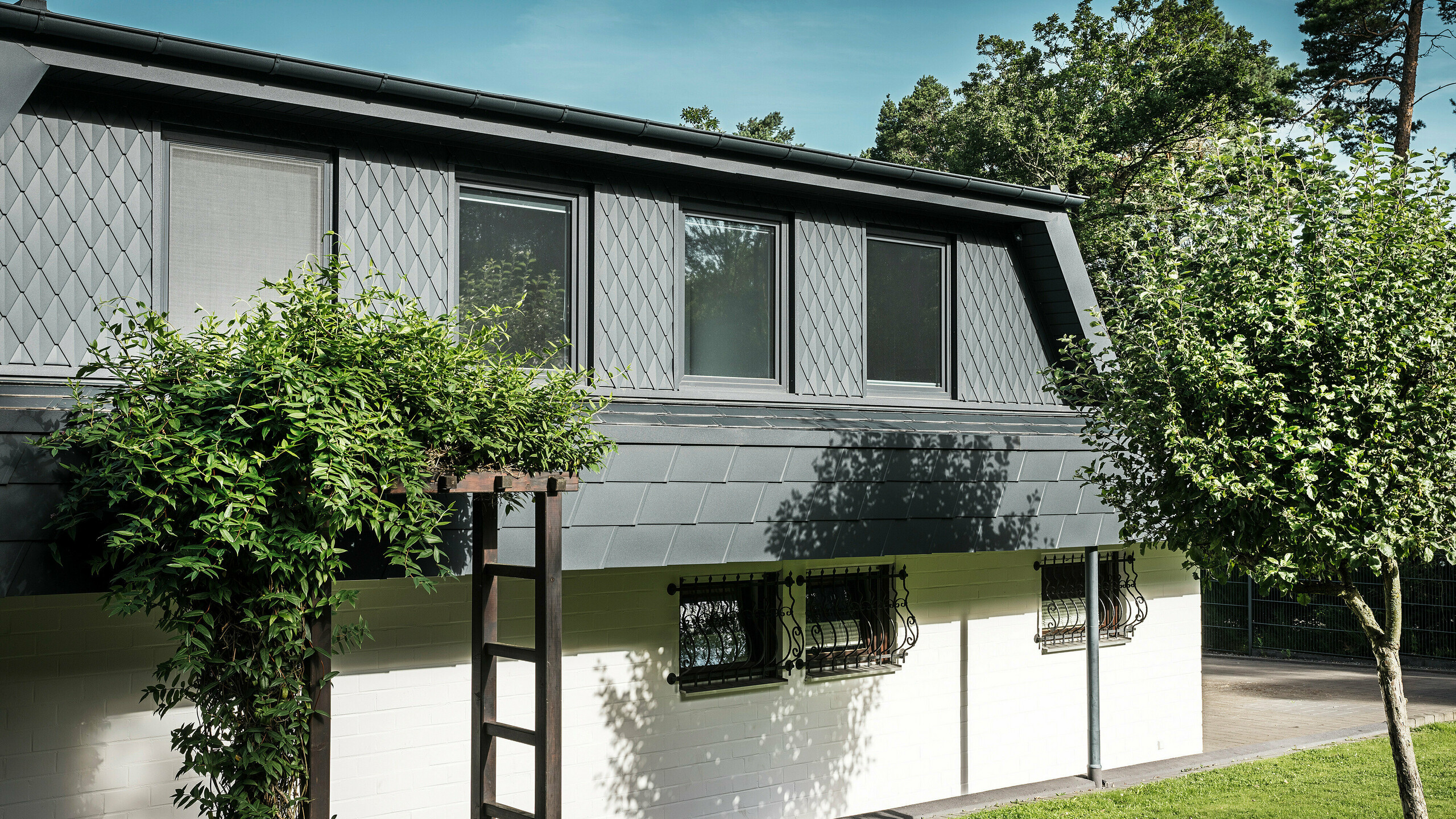 Bočný pohľad na rodinný dom v Mechernichu s veľkými lichobežníkovými vikiermi. Vikiere sú osadené malými strešnými šablónami PREFA v antracitovej farbe P.10, zatiaľ čo strecha je pokrytá osvedčenými šindľami PREFA DS.19, tiež v antracitovej farbe P.10. Hliníkové strešné šablóny a šindle predstavujú estetické a udržateľné riešenie strechy a fasády odolné voči poveternostným vplyvom. Tradičný rodinný dom je obklopený udržiavanou záhradou a množstvom stromov.