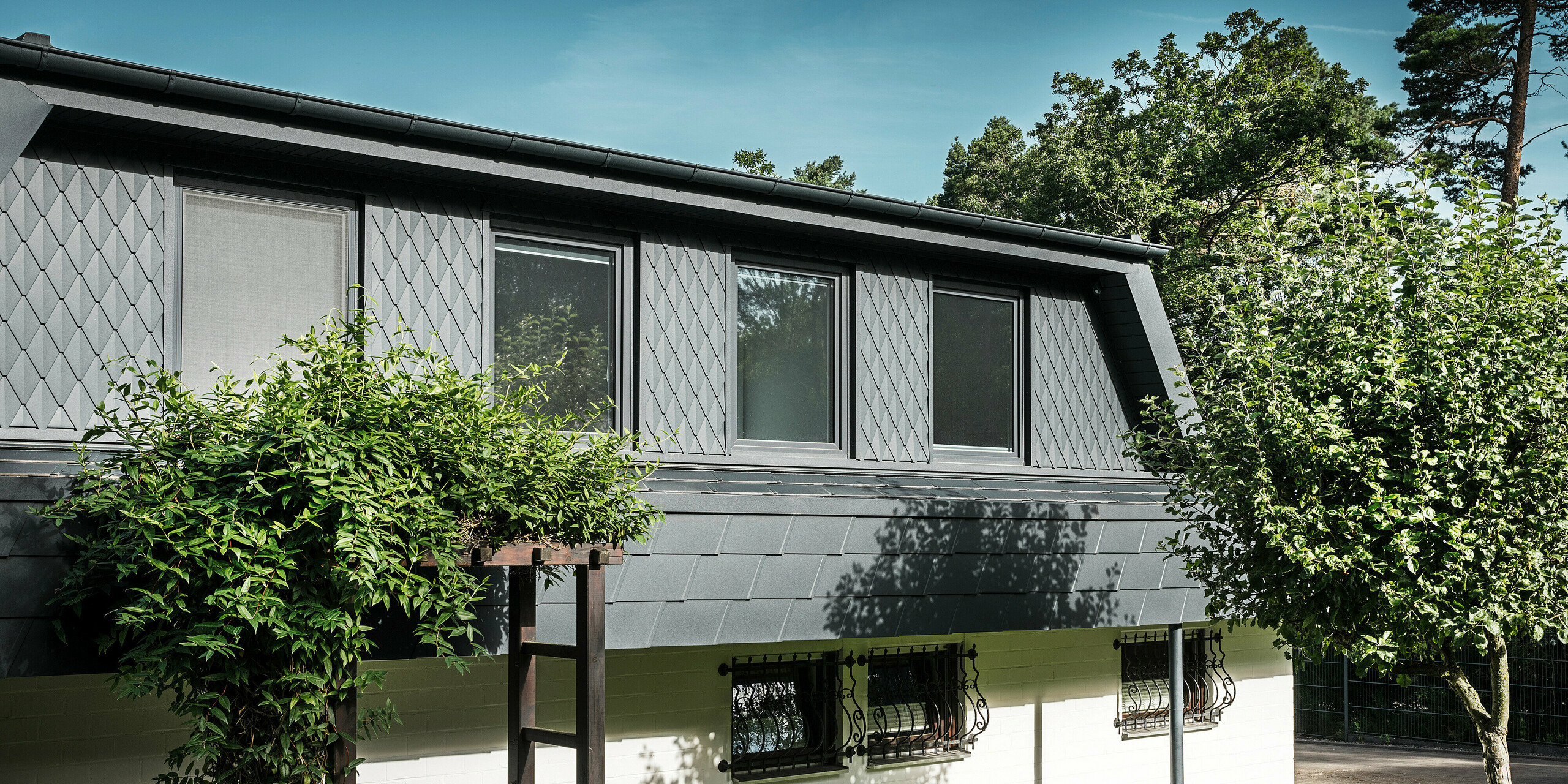 Bočný pohľad na rodinný dom v Mechernichu s veľkými lichobežníkovými vikiermi. Vikiere sú osadené malými strešnými šablónami PREFA v antracitovej farbe P.10, zatiaľ čo strecha je pokrytá osvedčenými šindľami PREFA DS.19, tiež v antracitovej farbe P.10. Hliníkové strešné šablóny a šindle predstavujú estetické a udržateľné riešenie strechy a fasády odolné voči poveternostným vplyvom. Tradičný rodinný dom je obklopený udržiavanou záhradou a množstvom stromov.