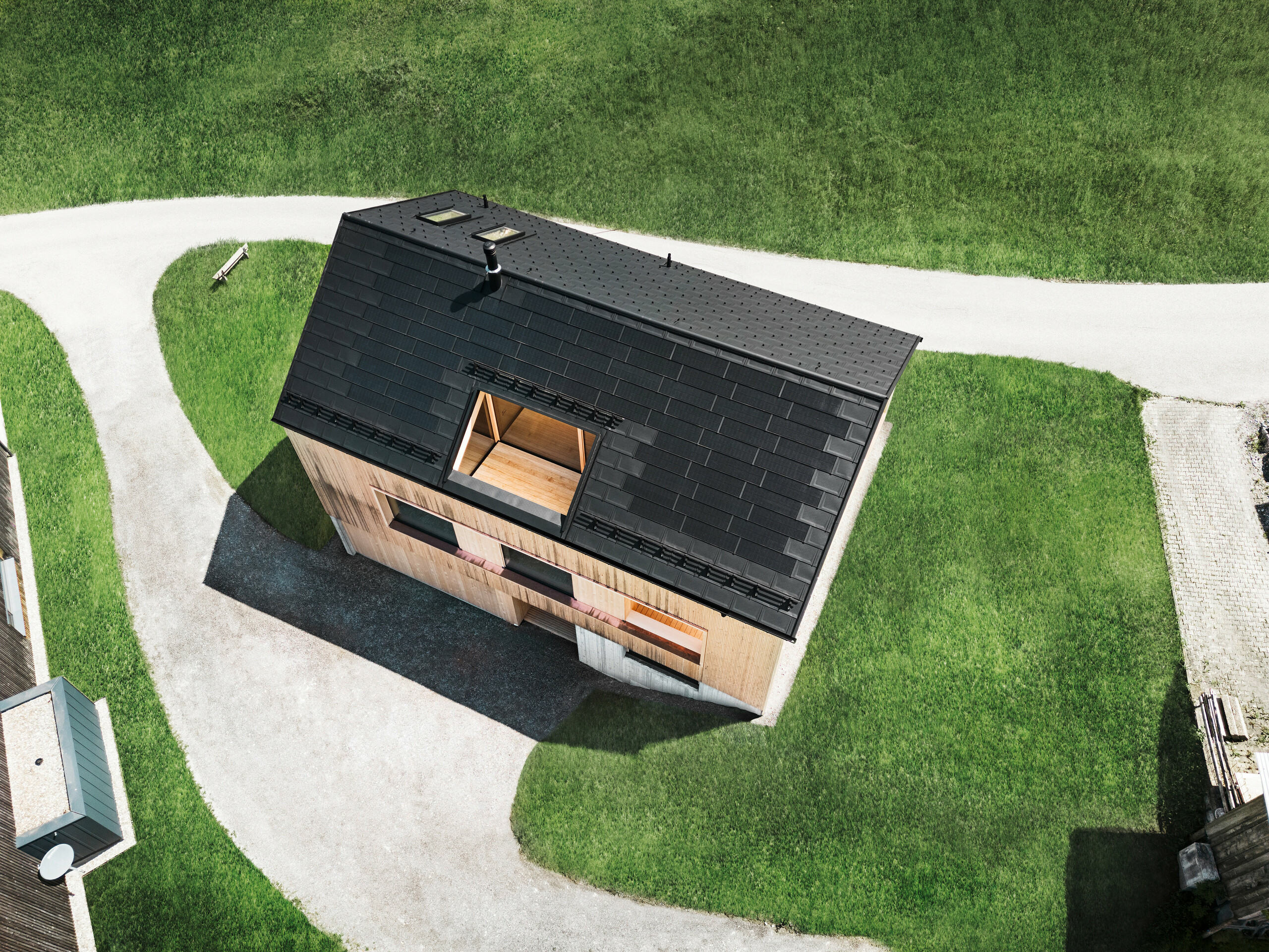 Záber z dronu s pohľadom zhora na ekologický rodinný dom v Eggu, ktorý sa odlišuje od okolitých zelených trávnikov svojou čiernou solárnou strechou PREFA a tradičnými strešnými panelmi PREFA R.16. Strategicky umiestnené solárne panely maximalizujú energetickú účinnosť, zatiaľ čo jasná geometria strechy a prirodzene vyzerajúca drevená fasáda vytvárajú spojenie medzi moderným bývaním a vidieckym prostredím. Tento dom je stelesnením progresívnej, ekologicky uvedomelej výstavby, ktorá je funkčne aj vizuálne pôsobivá.
