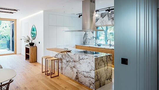 Záber zachytáva elegantný interiér budapeštianskej viacgeneračnej vily s modernou, otvorenou kuchyňou. Výraznými prvkami sú mramorované prírodné kamenné povrchy kuchynského ostrovčeka a zadnej steny v kombinácii s teplými dubovými akcentmi a kvalitným vstavaným nábytkom. Veľkorysé okenné plochy prepúšťajú množstvo denného svetla a harmonicky prepájajú priestor s okolitou zeleňou. Čisté línie, výber ušľachtilých materiálov a precízne spracovanie odrážajú vysoké architektonické nároky budovy, ktorej exteriér je doplnený trvácnymi hliníkovými produktmi PREFA.