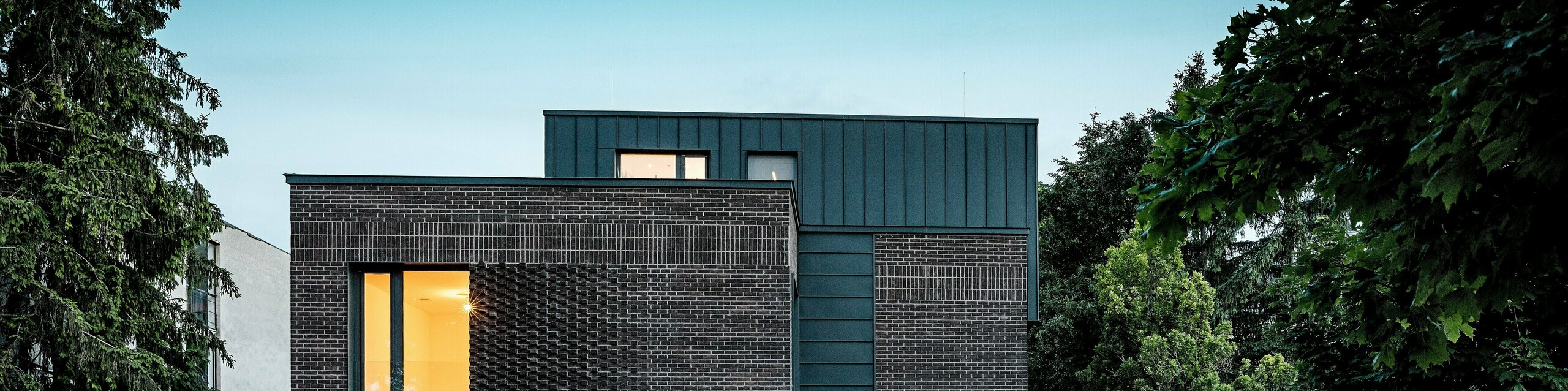 Na obrázku je moderný bytový dom v Budapešti, ktorého kubická strešná konštrukcia z hliníka PREFALZ v odtieni P.10 antracit vytvára výrazný kontrast s historickým okolím. Vertikálne uhlové stojaté falce vytvárajú zaujímavú hru svetla a zdôrazňujú jasný architektonický jazyk. Vysoko kvalitné hliníkové produkty PREFA dodávajú budove nielen elegantný, nadčasový vzhľad, ale presviedčajú aj svojou dlhou životnosťou, odolnosťou voči poveternostným vplyvom a precíznou spracovanosťou. Antracitová farba fasády a strechy pôsobí ako silný magnet pre pohľad a podčiarkuje inovatívny charakter stavby.