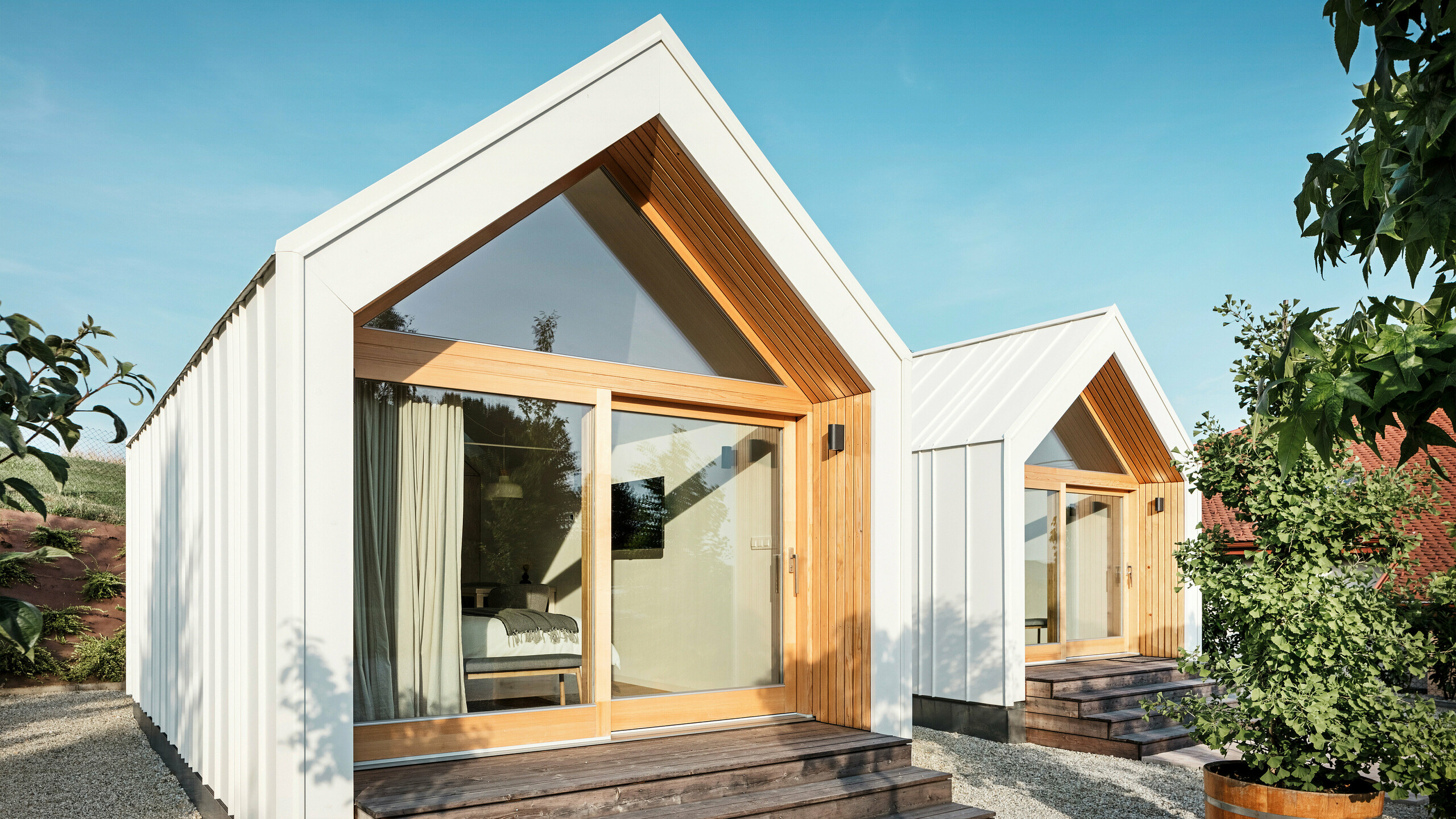 Pohľad spredu na dva malé domčeky ubytovacieho zariadenia „Pri Momi“ v Kančevci v Slovinsku s veľkými presklenými priečeliami, navrhnuté so strechou PREFALZ a fasádnym obkladom z čistej bielej farby P.10 a prírodného dreva. Minimalistické mikrodomčeky majú široké drevené schody vedúce ku vchodom a ponúkajú priamy pohľad do útulných interiérov. Čisté línie a kombinácia hliníka a dreva podčiarkujú moderný a prívetivý dizajn rekreačného ubytovania.