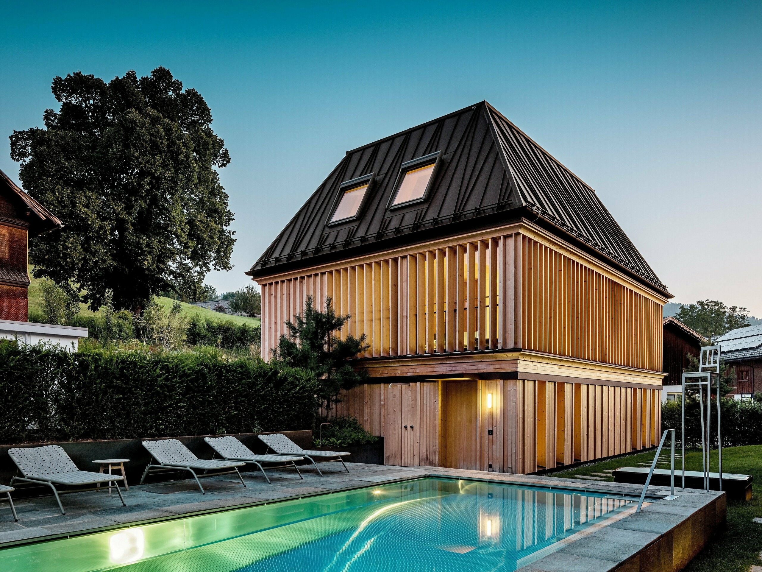 „Kúpeľný dom“ hotela Hirschen v Schwarzenbergu za súmraku, s výhľadom na osvetlený vonkajší bazén v popredí. Budova zaujme svojou modernou valbovou strechou z PREFALZ vo farbe P.10 orieškovohnedá s presne vyhotovenými, čiastočne šikmými stojatými falcami. Dve strešné okná zabezpečujú denné svetlo vo vnútri. Fasáda sa skladá z vertikálnych drevených lamiel po celej výške poschodia, ktoré dodávajú budove štruktúru a ľahkosť. V spodnej časti dominuje prírodné drevo. Obklopený upravenou zeleňou a zasadený do kopcovitej krajiny Bregenzerwaldu vytvára harmonické spojenie architektúry, prírody a oddychu.