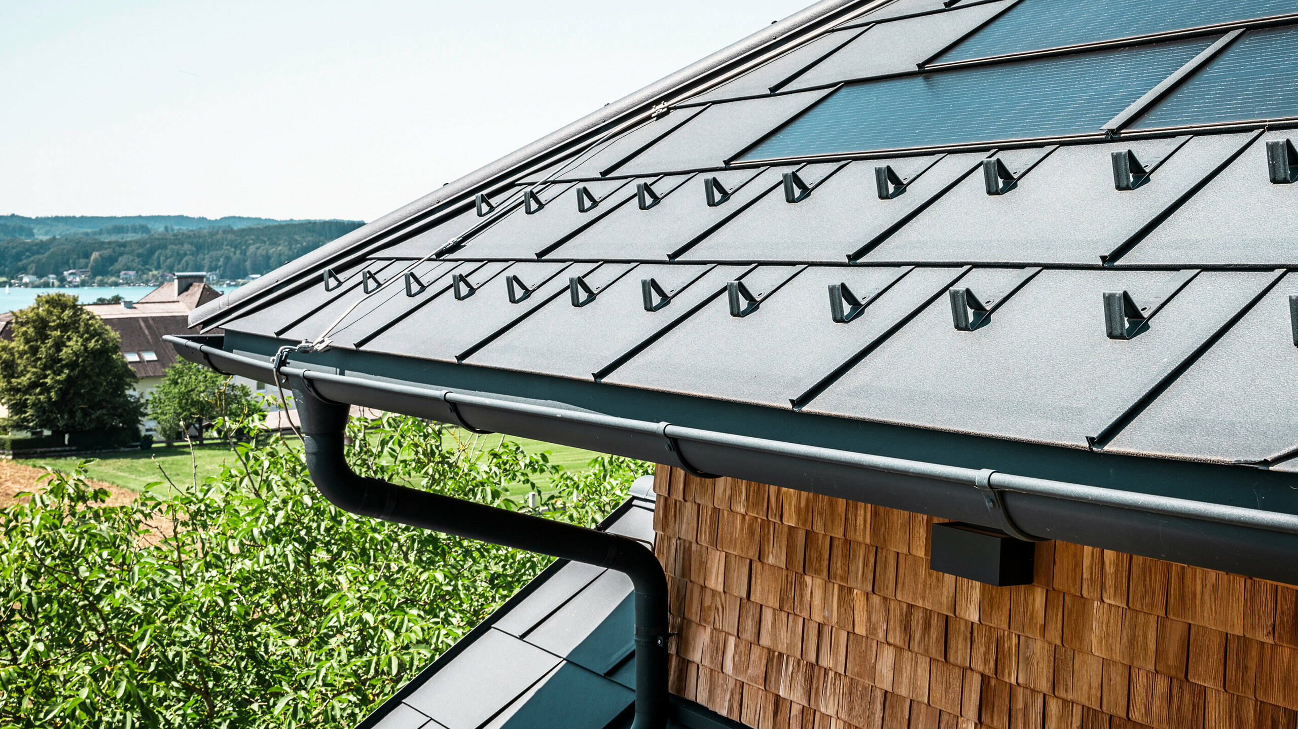 Zenz'n Stub'n v Schörflingu pri Attersee predstavuje novo zrekonštruovanú strechu s inovatívnymi solárnymi panelmi PREFA a odolnými strešnými panelmi PREFA R.16 vo farbe P.10 antracitová. Štýlová hliníková strecha spája moderný dizajn s udržateľnou výrobou energie. Aj odvodnenie strechy, pozostávajúce z odvodňovacej rúry a polkruhového žľabu vo farbe P.10 antracit, harmonicky zapadá do celkového obrazu a podčiarkuje kvalitné spracovanie. V pozadí sa leskne modrá farba jazera Attersee.